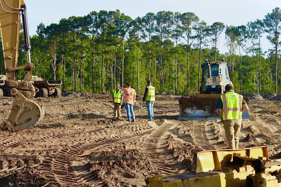 Construction site with workers in safety vests operating heavy machinery and inspecting the area, surrounded by trees.