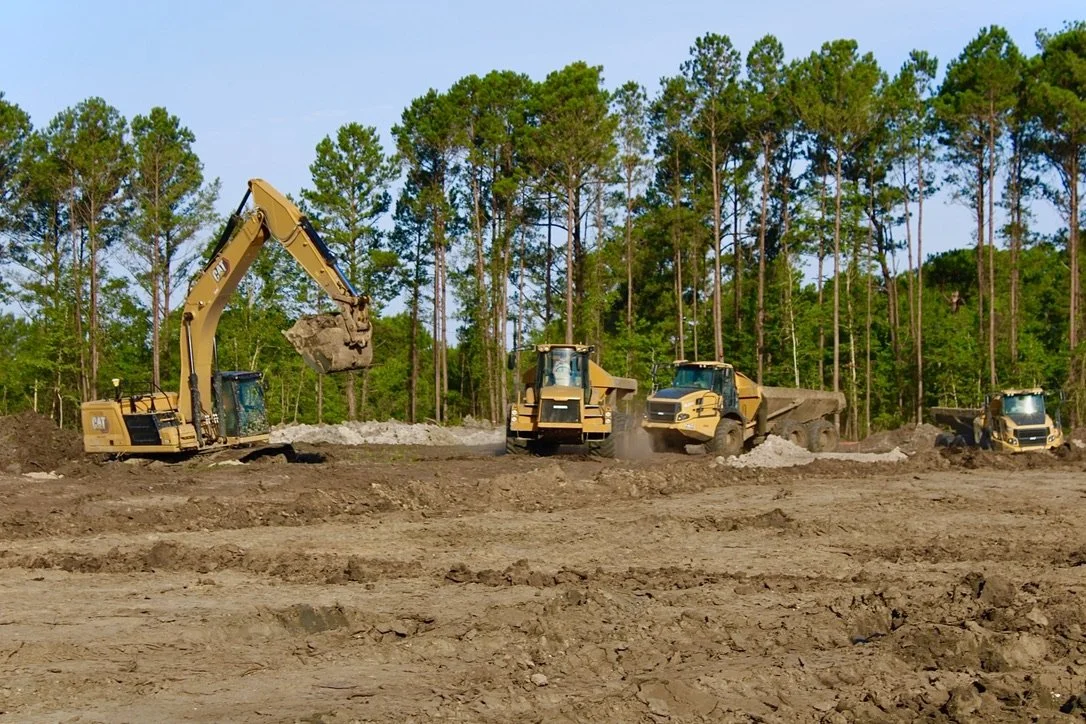 Construction site with excavators, bulldozers, and dump trucks working on clearing and leveling land surrounded by a forest of tall green pine trees.