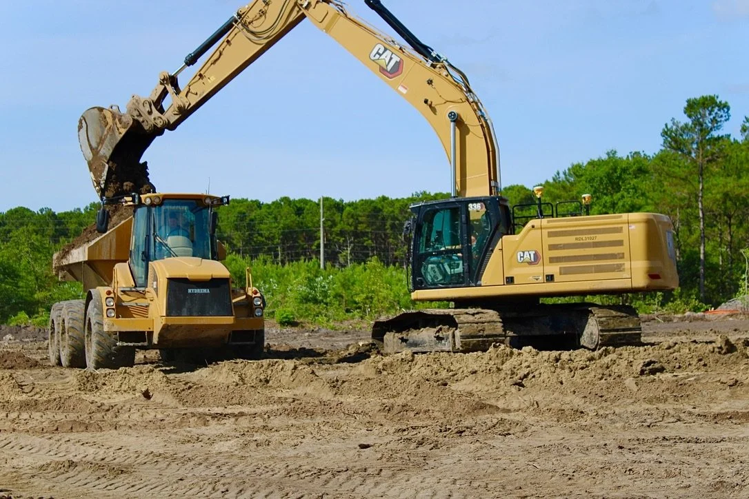 A construction site with a large excavator and a dump truck working together, against a backdrop of green trees and blue sky.