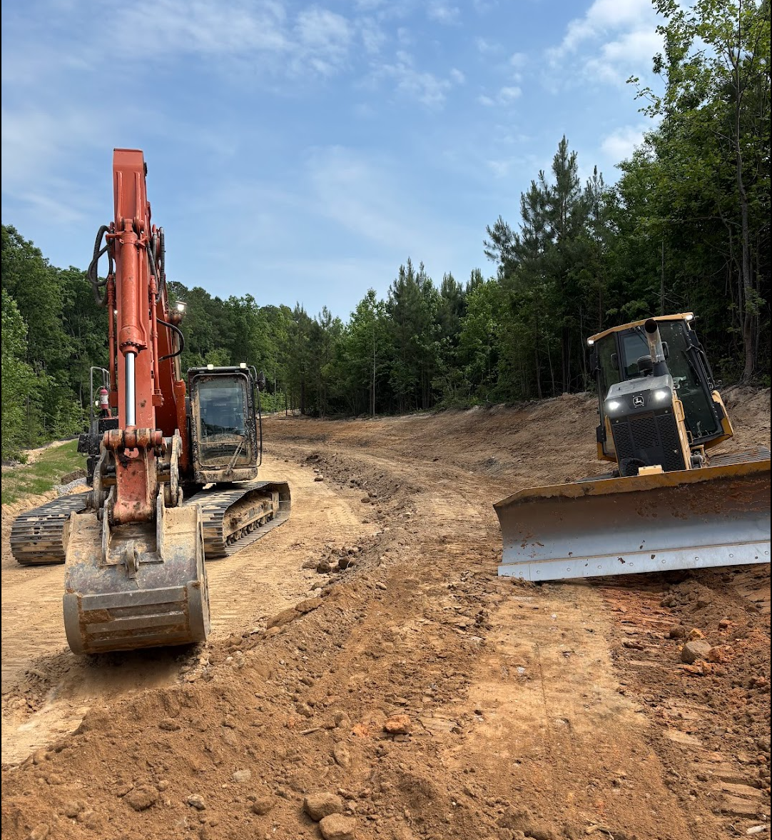 Two construction vehicles, a excavator and a bulldozer, working on a dirt road in a forested area with green trees and a partly cloudy blue sky.
