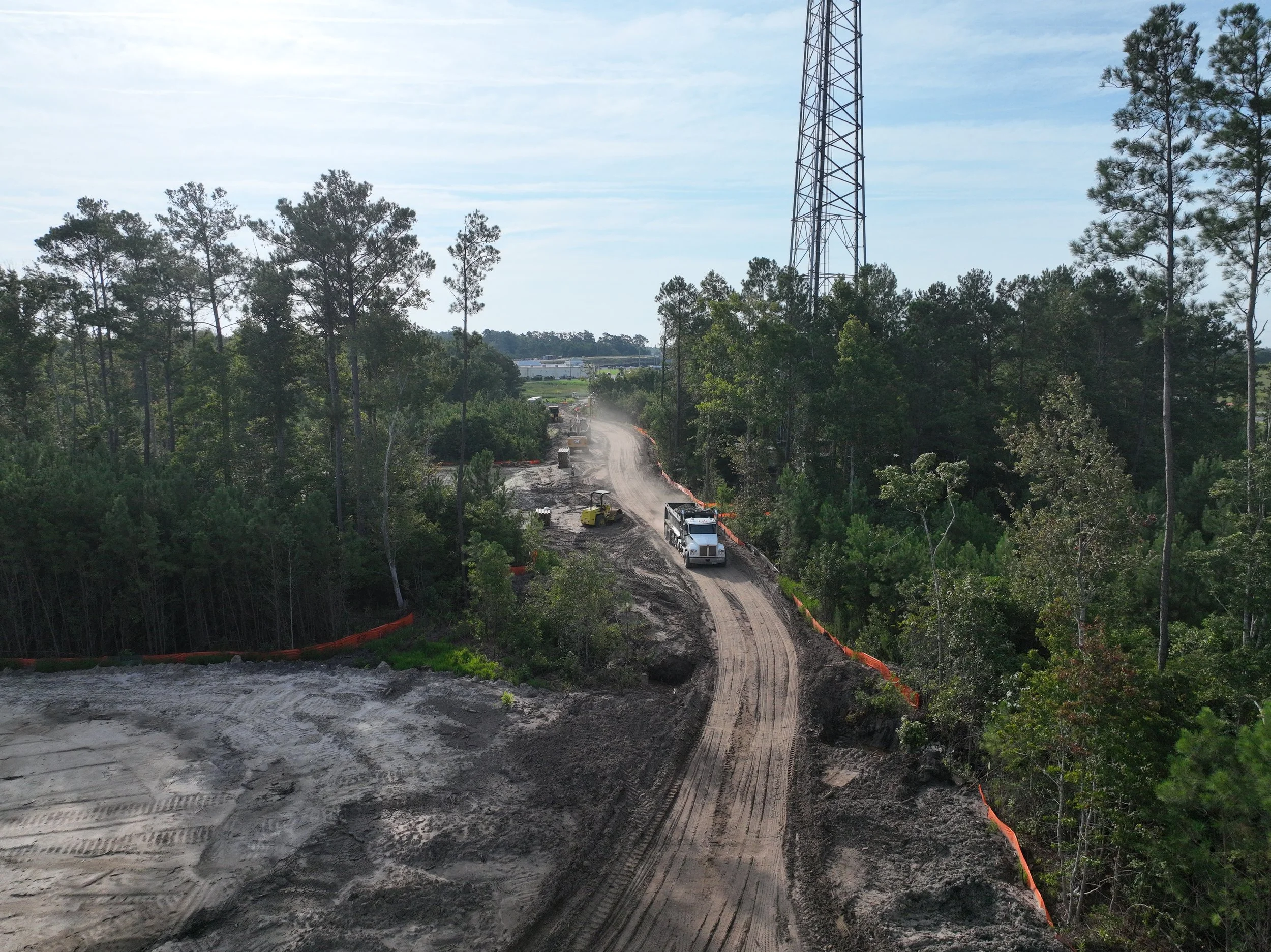 A construction site on a forested area with a dirt road, construction vehicles, and a tall communication tower in the background.