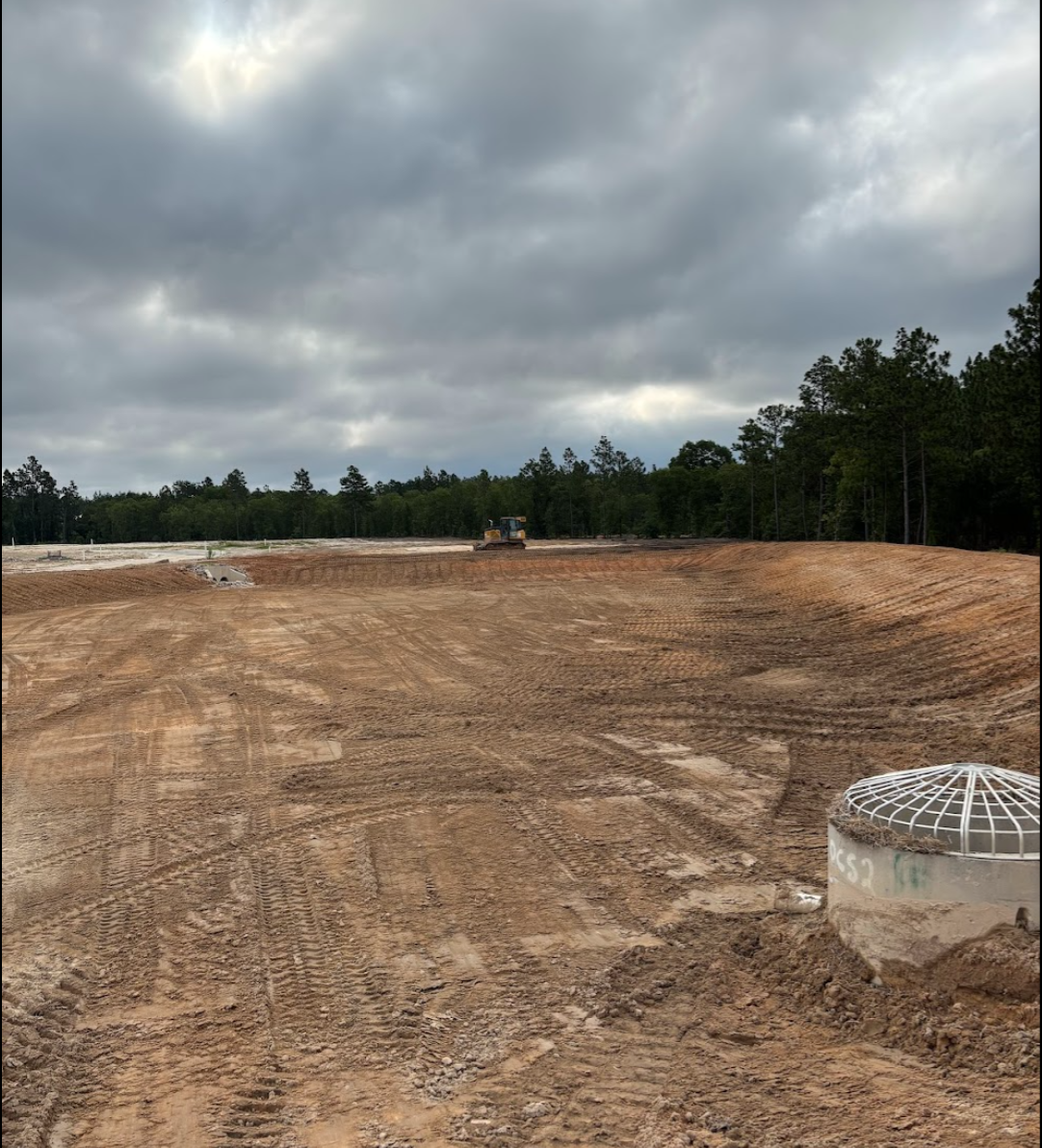 A construction site with disturbed dirt and a bulldozer in the background under a cloudy sky, with trees in the distance.