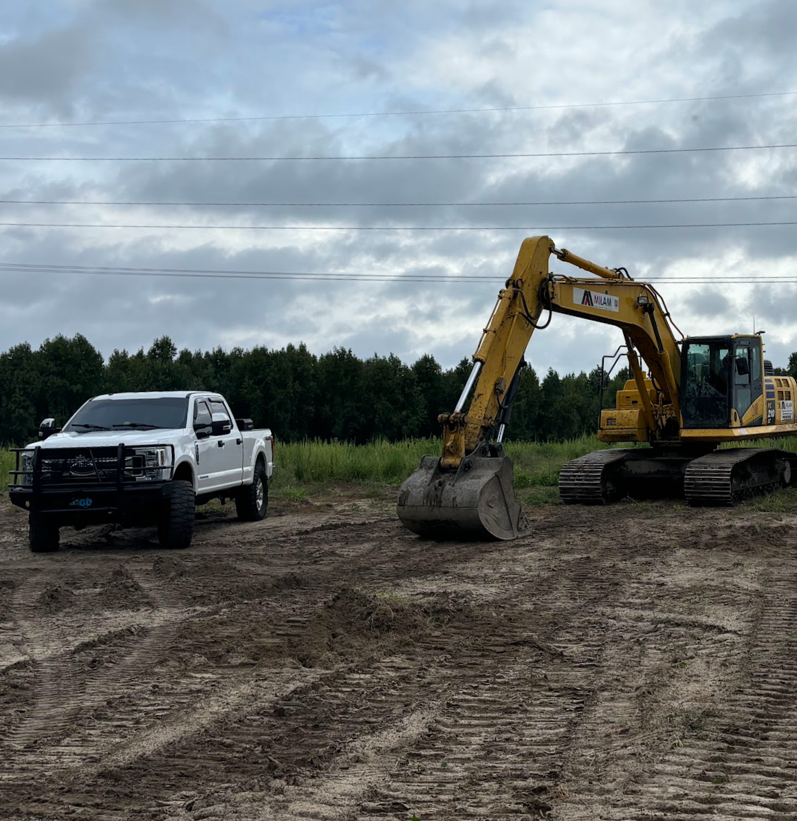 A construction site with a white pickup truck and a yellow excavator on dirt ground, with trees and cloudy sky in the background.
