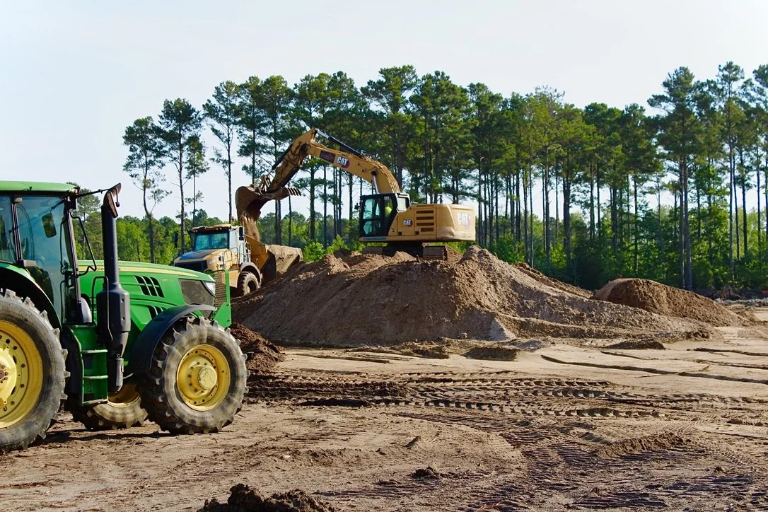 Construction site with green tractor in foreground, yellow excavator on dirt mounds, and forest in the background.