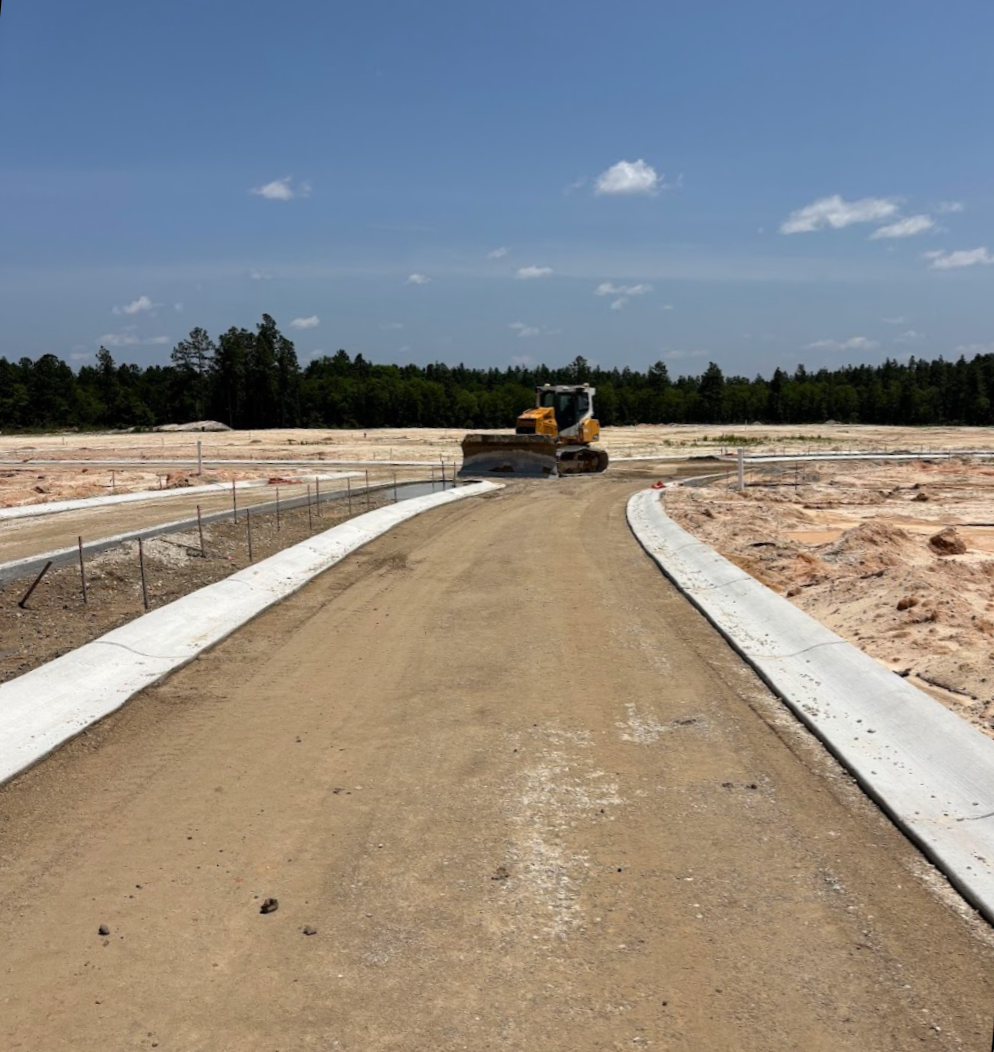 Construction site with a bulldozer working on a new road, dirt and gravel surface, blue sky with a few clouds, and trees in the background.