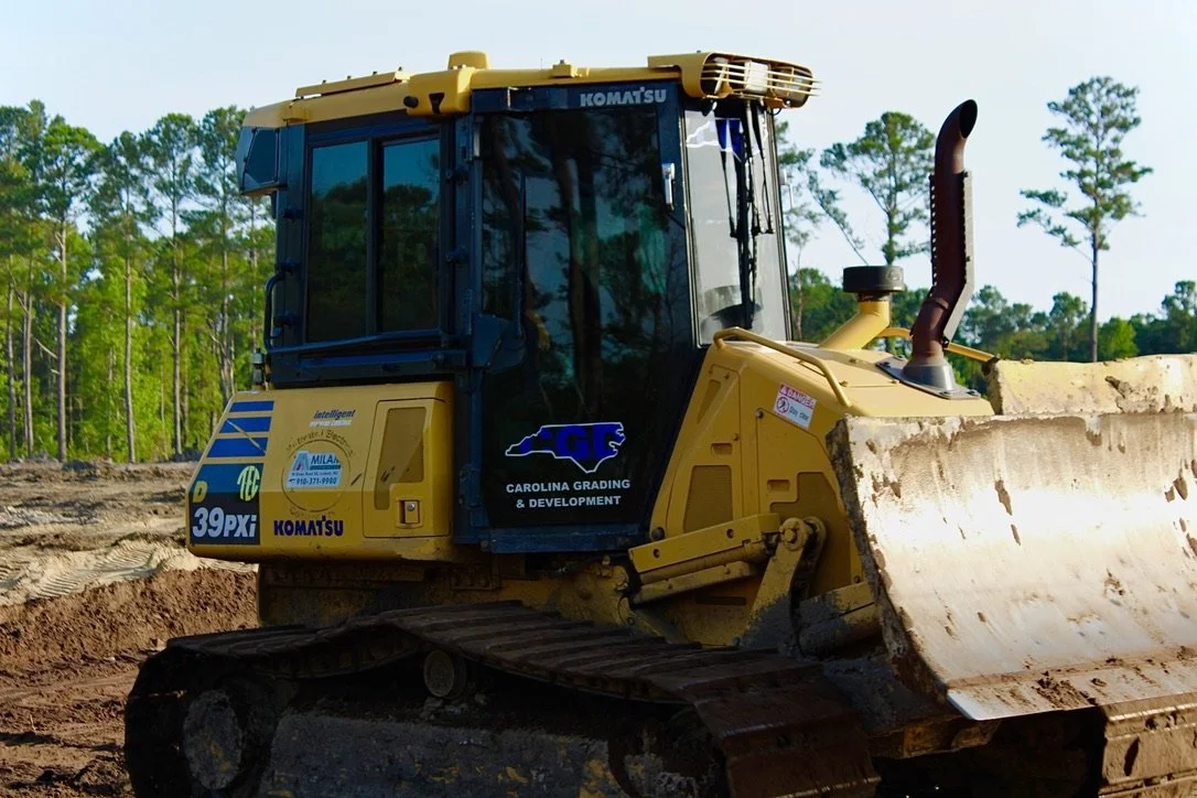 Yellow bulldozer on dirt construction site with trees in the background.