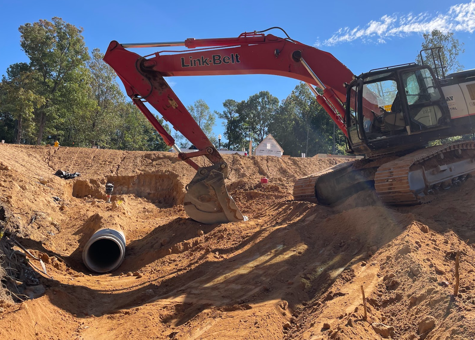 A red excavator labeled 'Link-Belt' working on a construction site, digging through sand and soil, with trees and a blue sky in the background.
