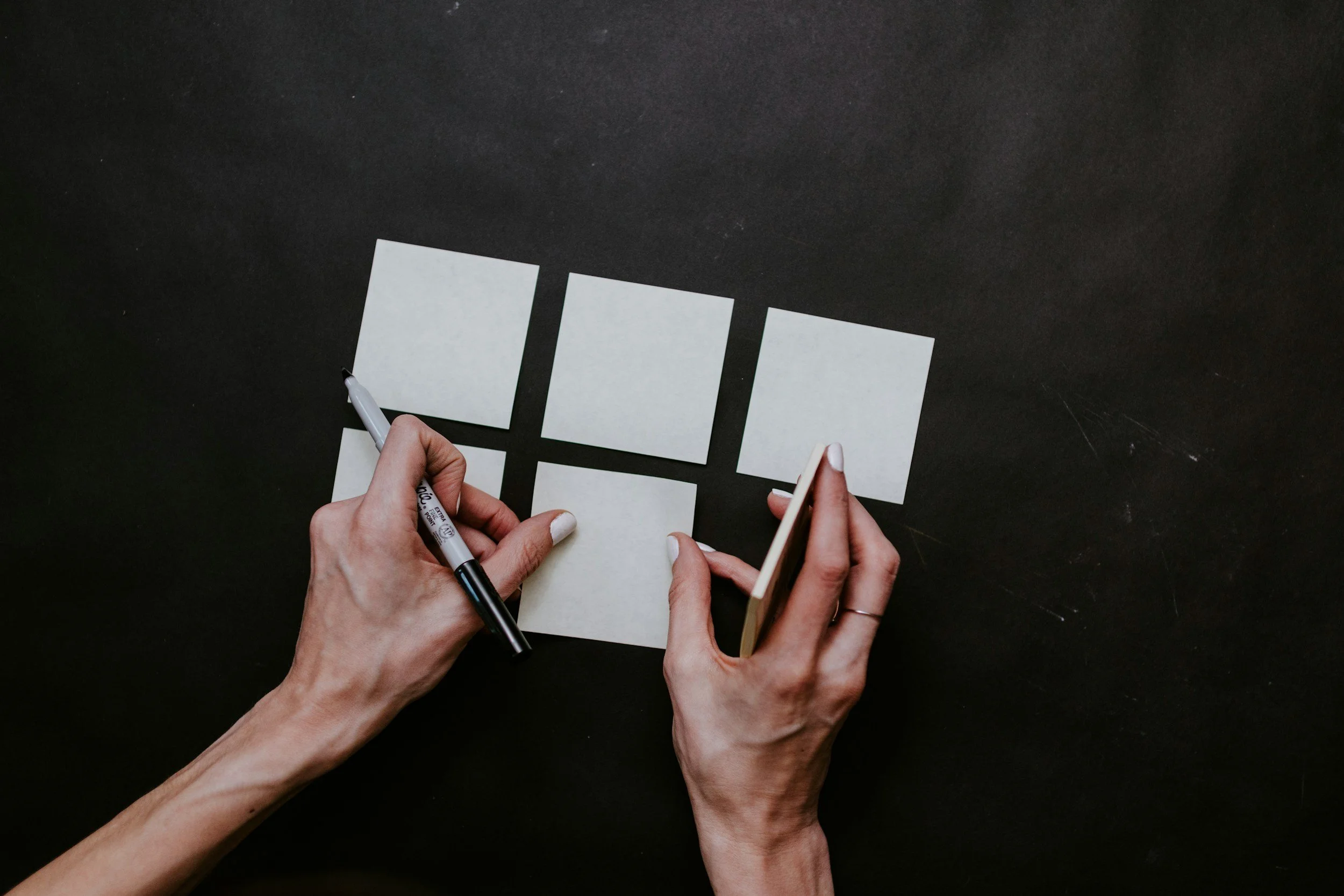 Hands writing on blank sticky notes arranged on a black surface.