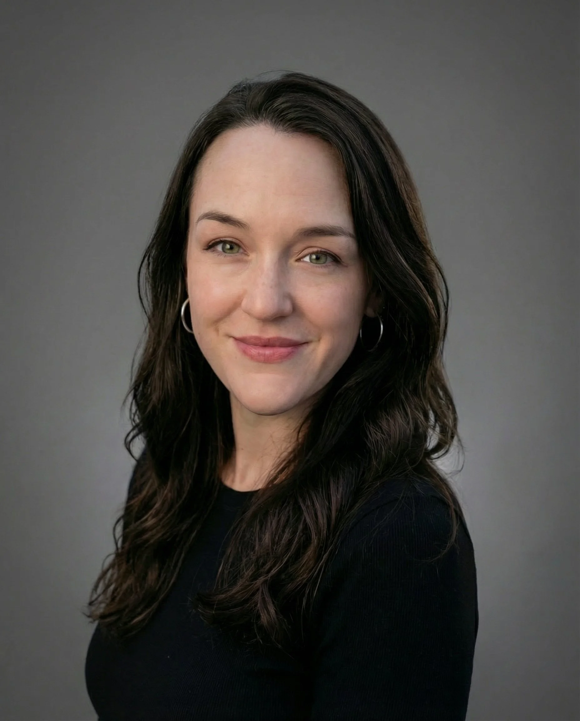 A woman with long dark hair, wearing a black top and silver hoop earrings, smiling at the camera against a plain gray background.