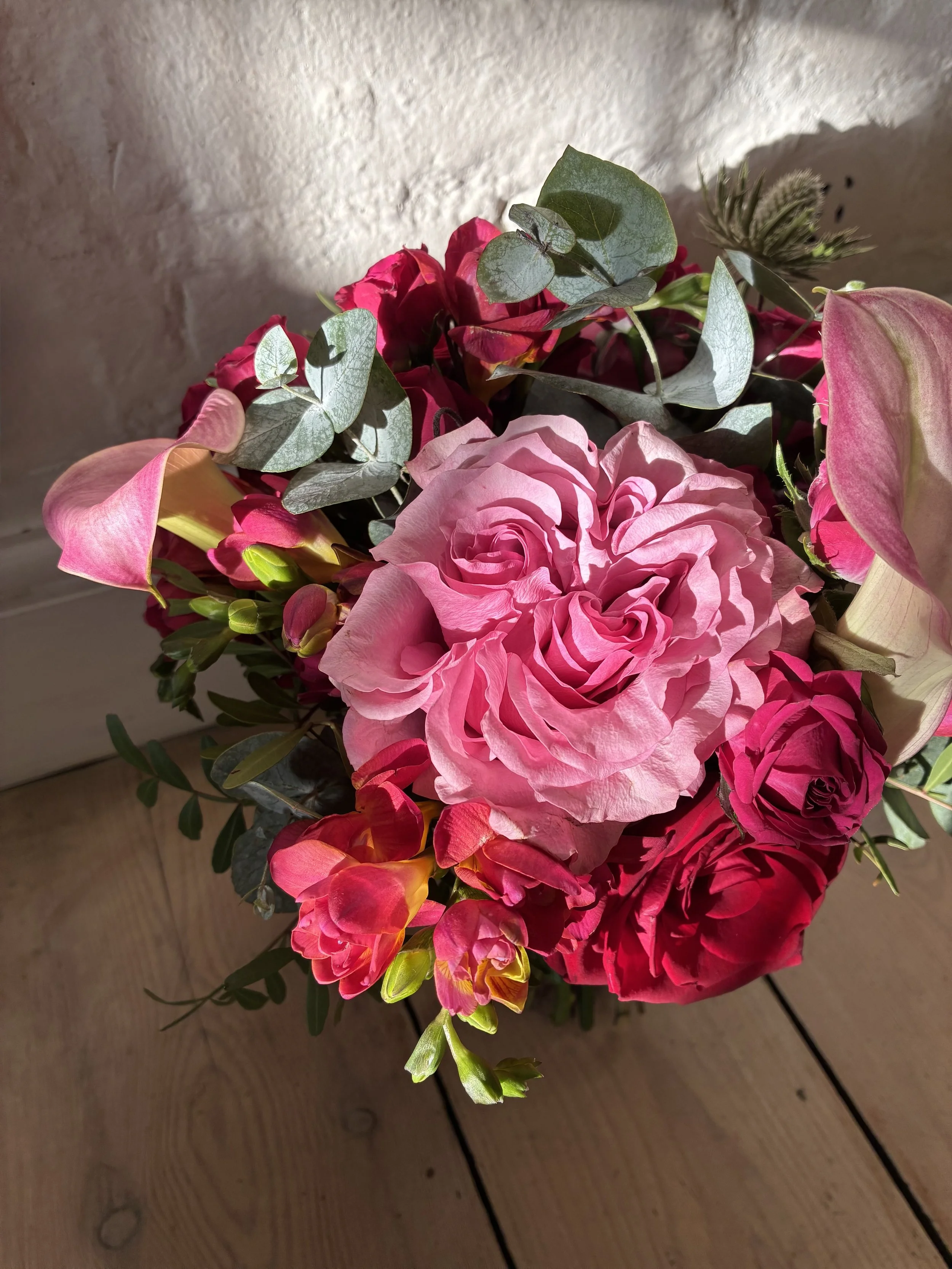 A bouquet of pink roses, red and pink calla lilies, and green foliage on a wooden surface.