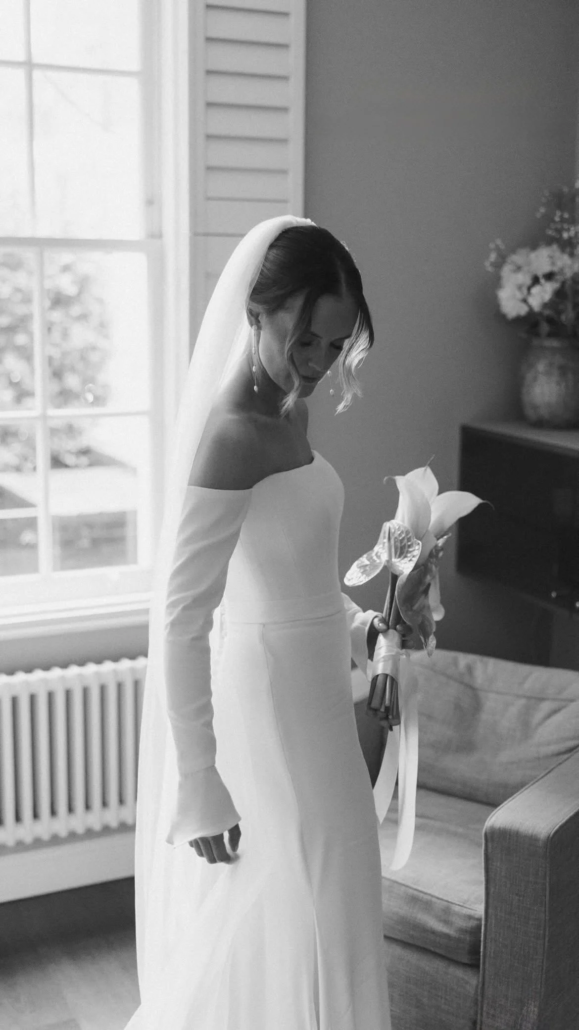A bride in an off-the-shoulder wedding gown holding a bouquet of flowers, standing indoors near a window and a sofa, with a vase of flowers in the background, depicted in black and white.