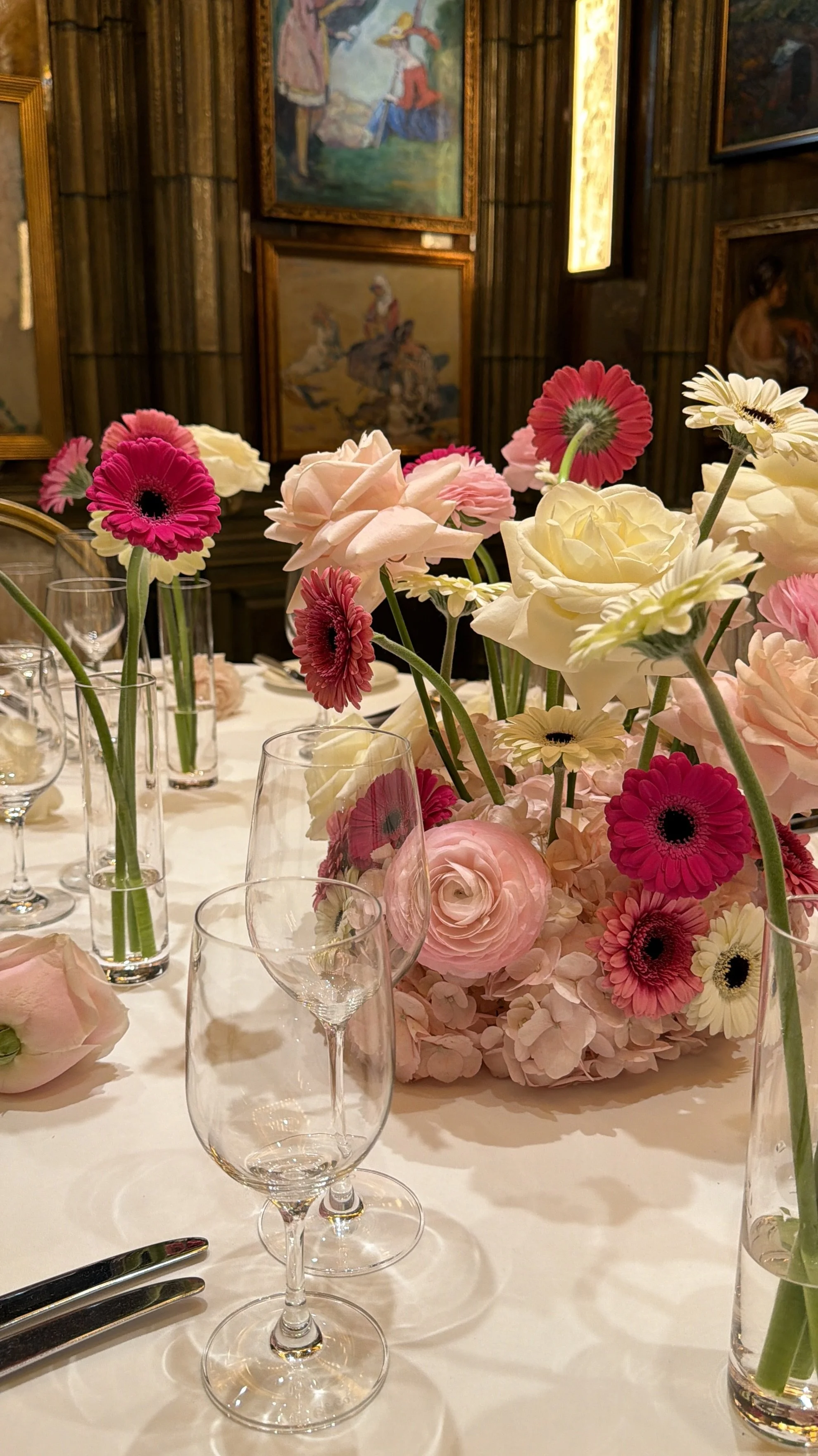 A table set with glassware and a floral centerpiece of pink, white, and cream flowers, with paintings hanging on a wooden-paneled wall in the background.