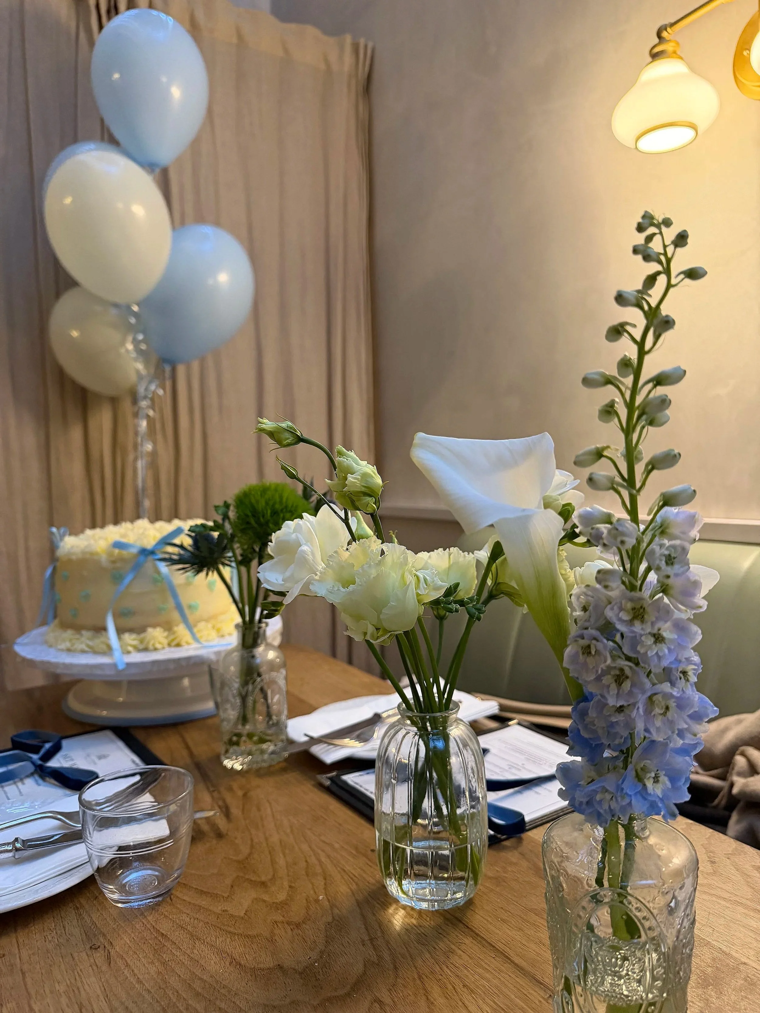 Decorative floral arrangement with white and light purple flowers in glass vases on a wooden table, nearby a birthday cake with a blue bow, and balloons in white and light blue in the background, decorated for a celebration.