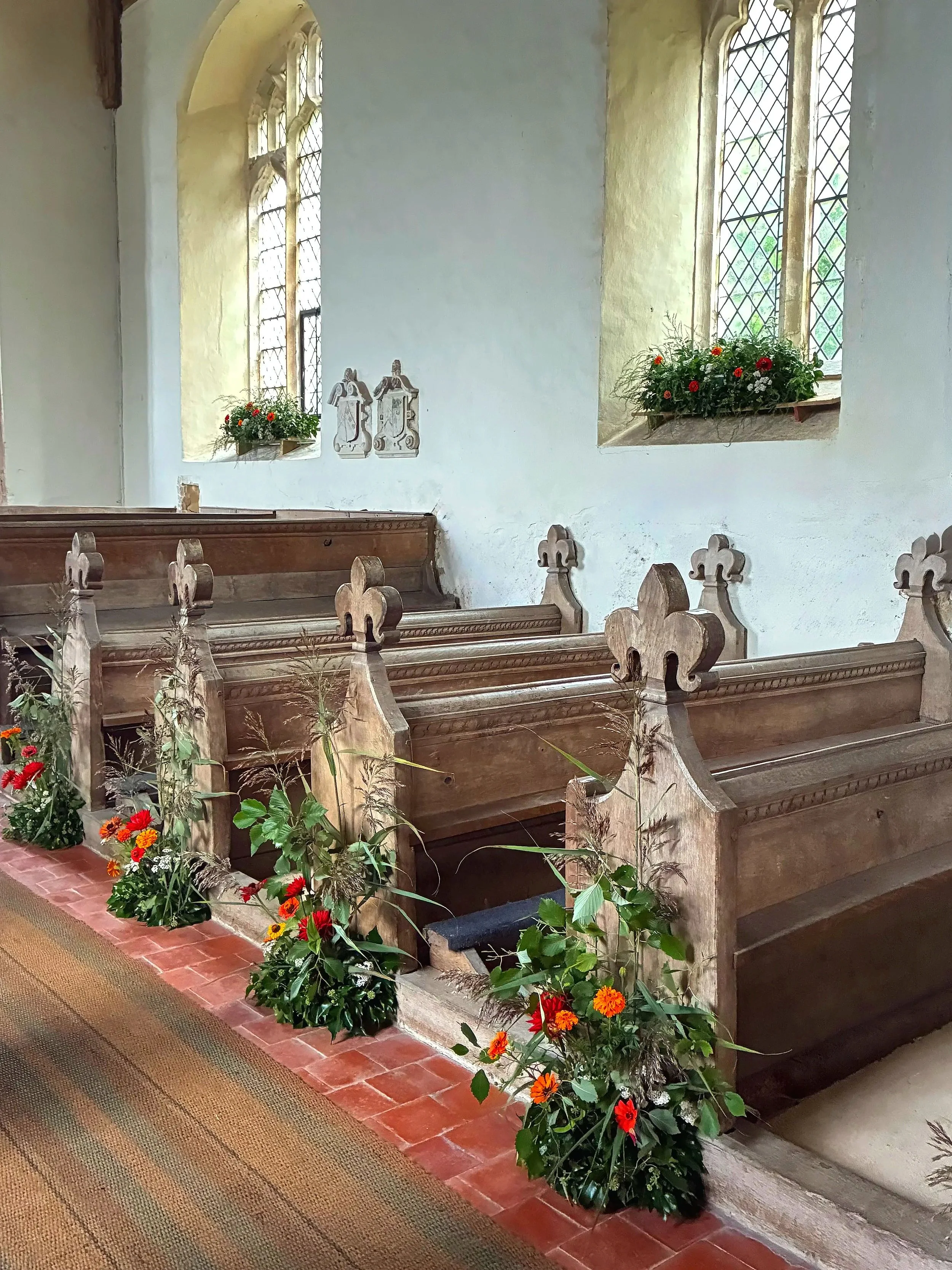 Inside an old church with wooden pews decorated with flowers, brick flooring, and stained glass windows with flower boxes.