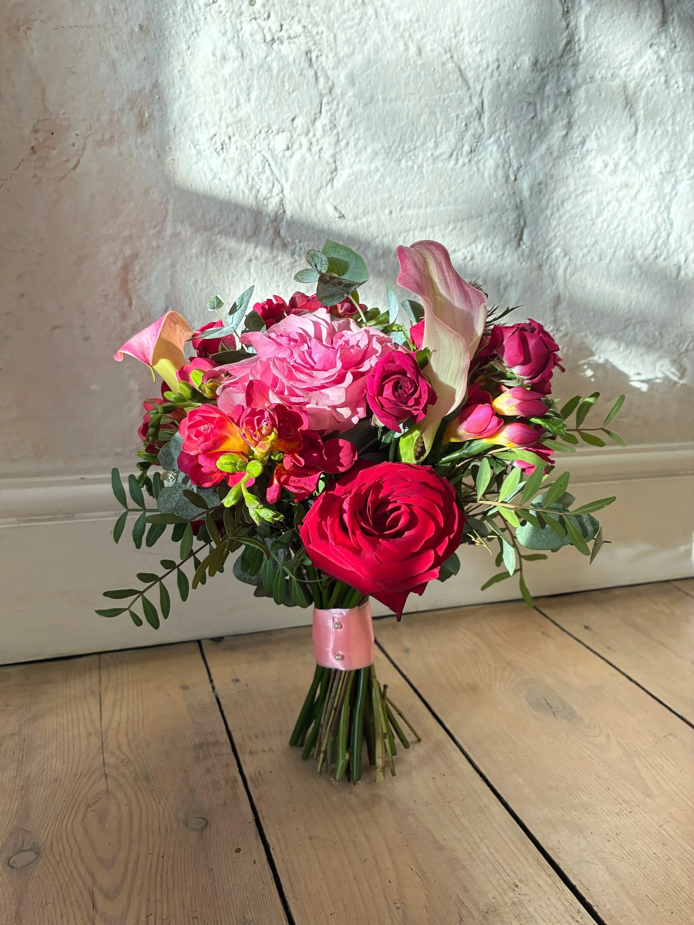 A bouquet of mixed pink and red flowers including roses, calla lilies, and small buds, tied with a pink ribbon, placed on a wooden floor with a textured white wall background.