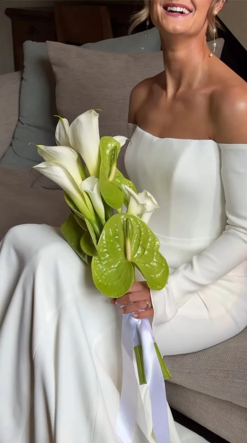 A woman in a white off-the-shoulder dress is sitting on a beige couch, smiling, and holding a bouquet of white calla lilies and green peace lilies.