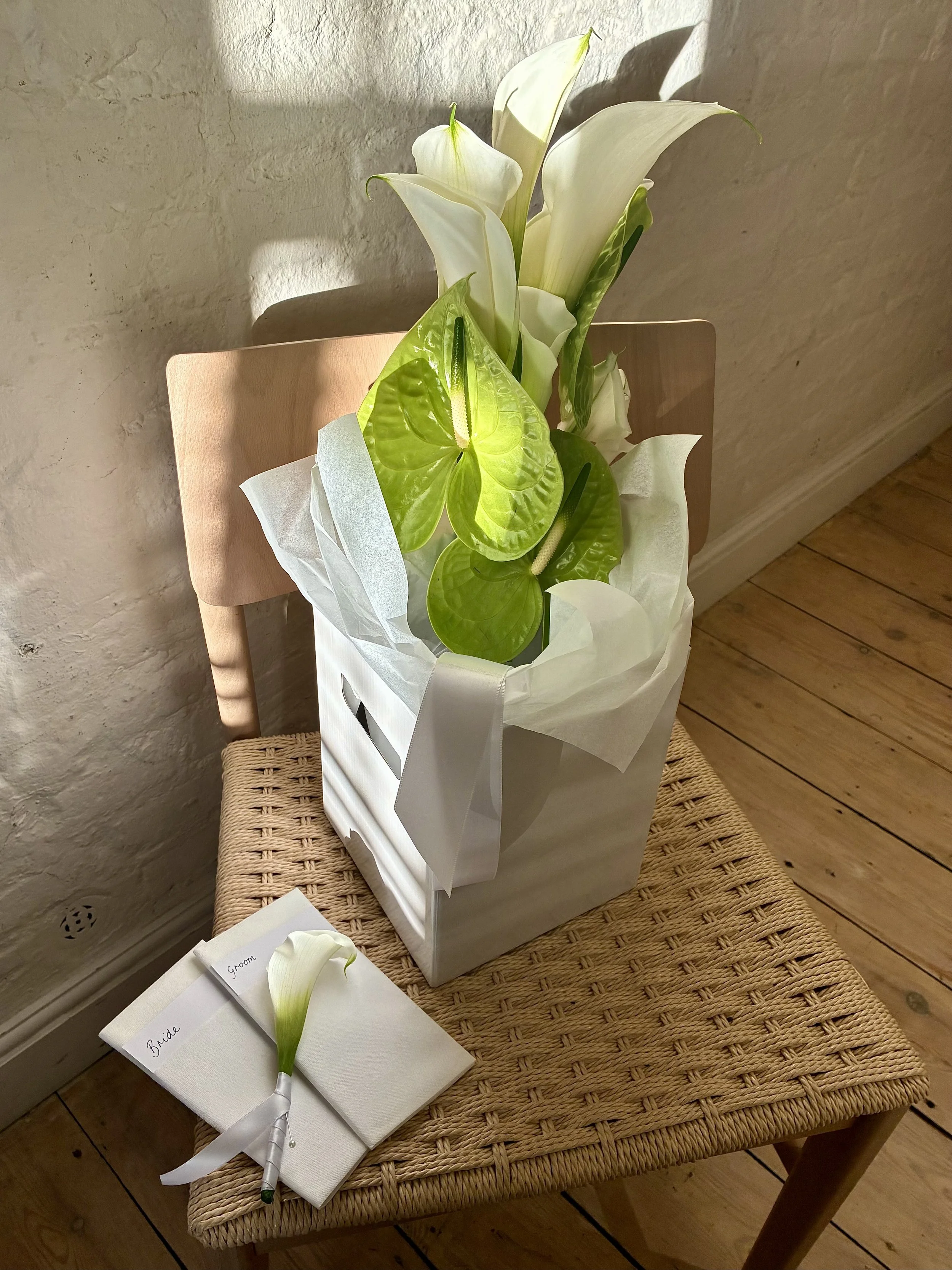A bouquet of white calla lilies and green anthuriums in a white box, placed on a chair in a room with wooden flooring and a textured wall. There's a white napkin with a calla lily and labels for "Bride" and "Groom" on the table.
