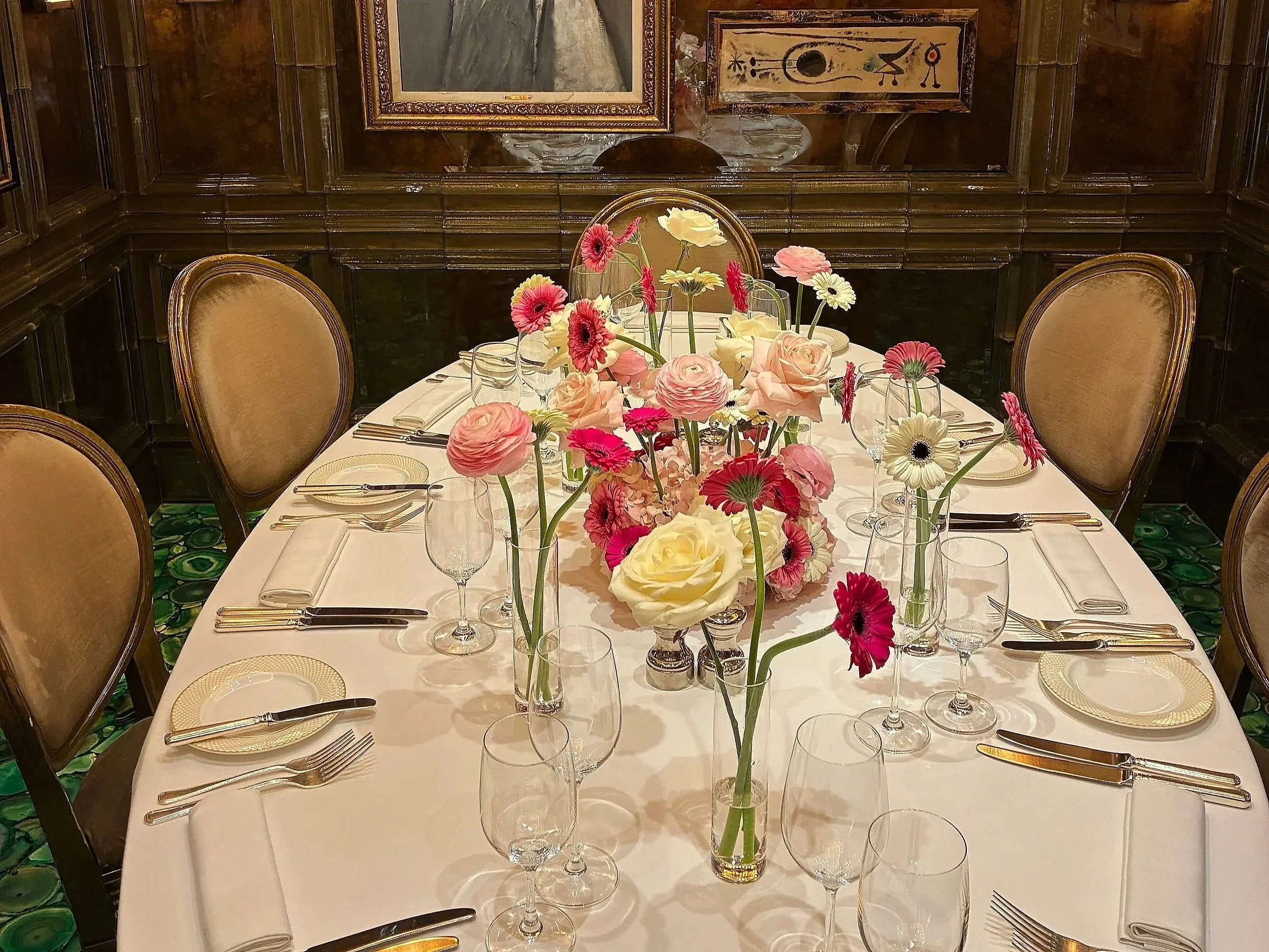 A round dining table with a white tablecloth, set with plates, silverware, glasses, and cloth napkins, with a centerpiece of pink, white, and cream flowers in vases, in a room with dark wood-paneled walls and framed artwork.