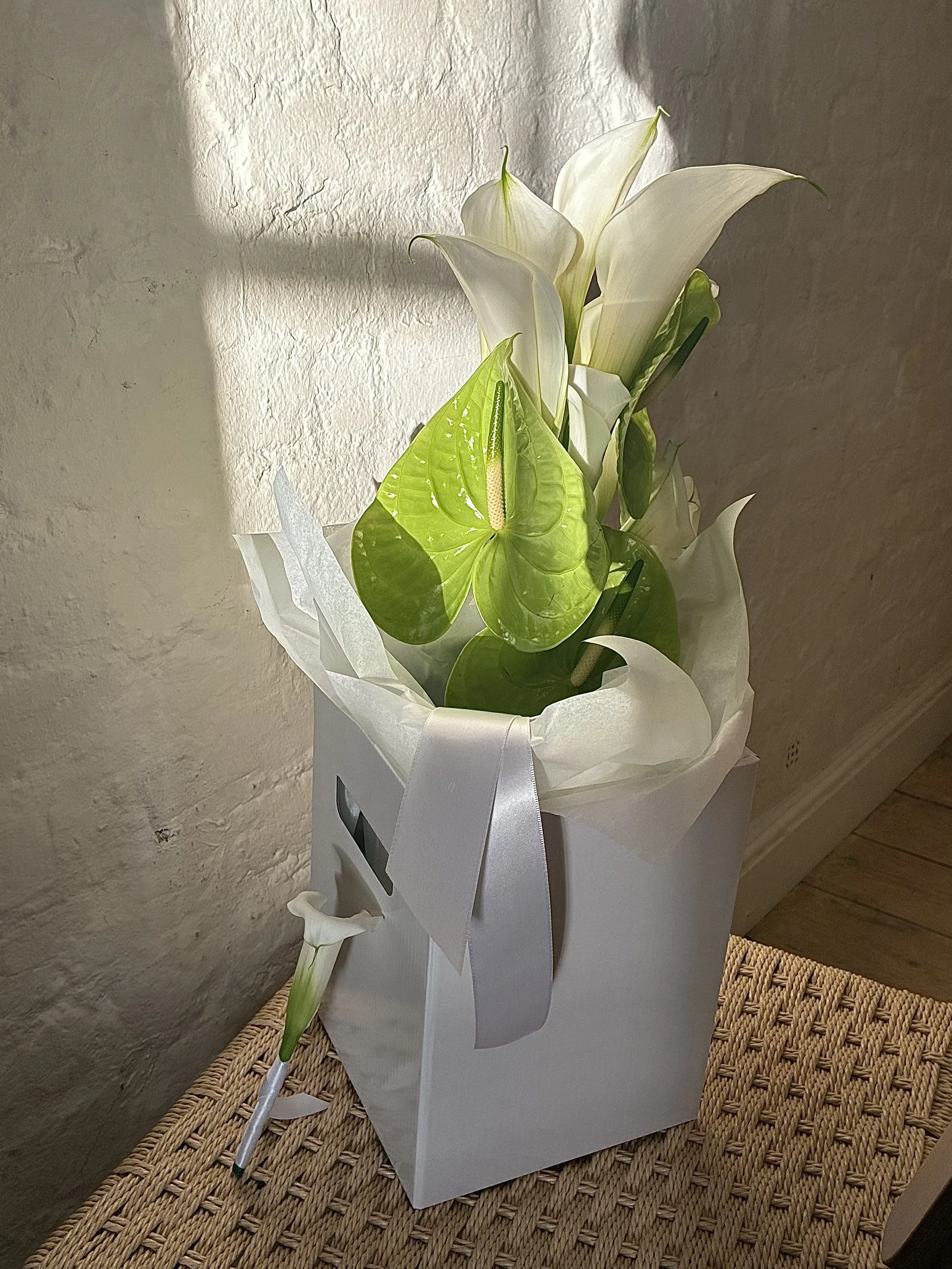 White gift box with a bouquet of white calla lilies and green anthurium flowers, decorated with white tissue paper and a white ribbon, placed on a woven table.