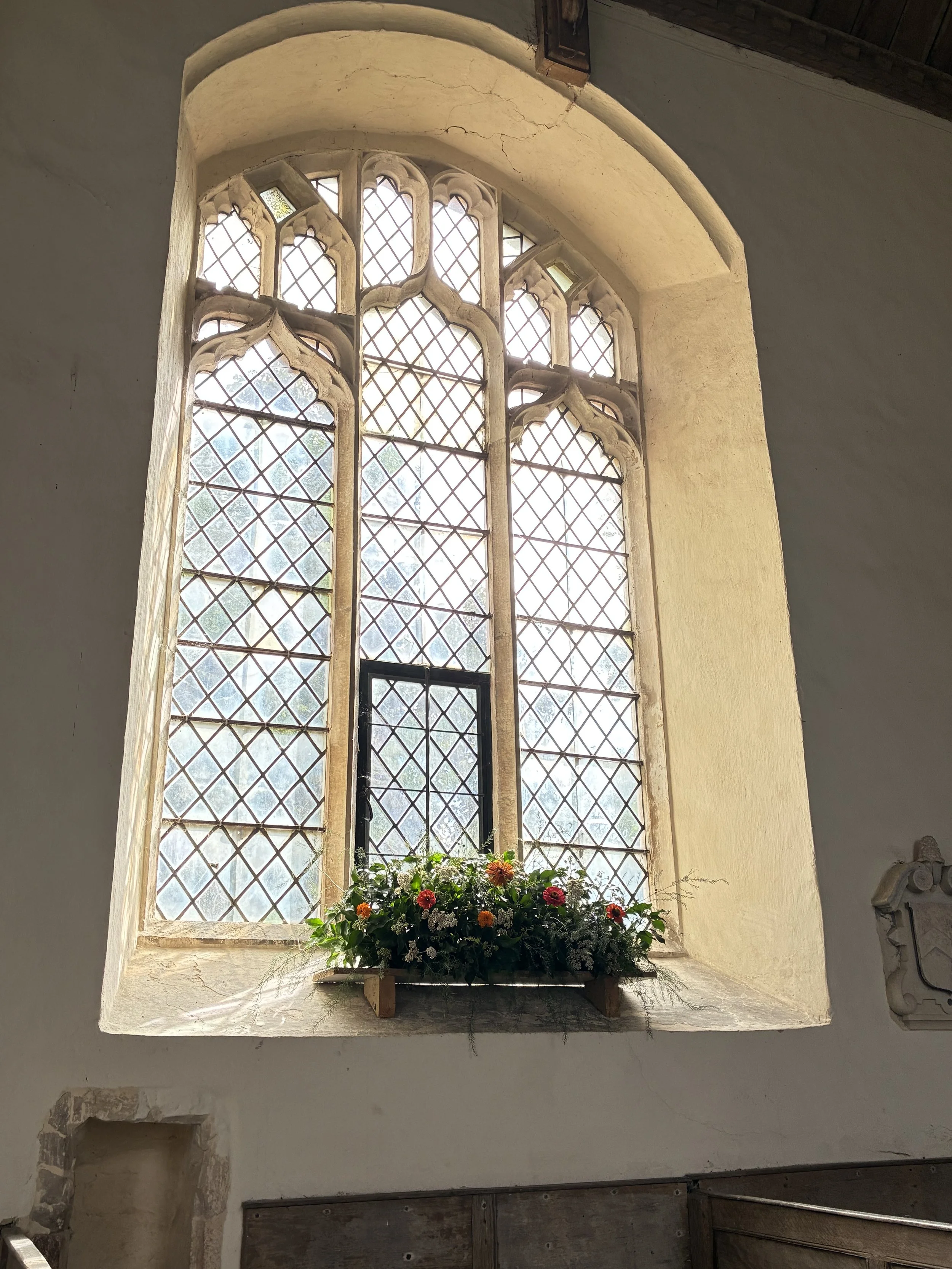 A large Gothic-style stained glass window with intricate diamond-shaped glass panes and decorative stone tracery, with a flower box containing colorful flowers on the windowsill.