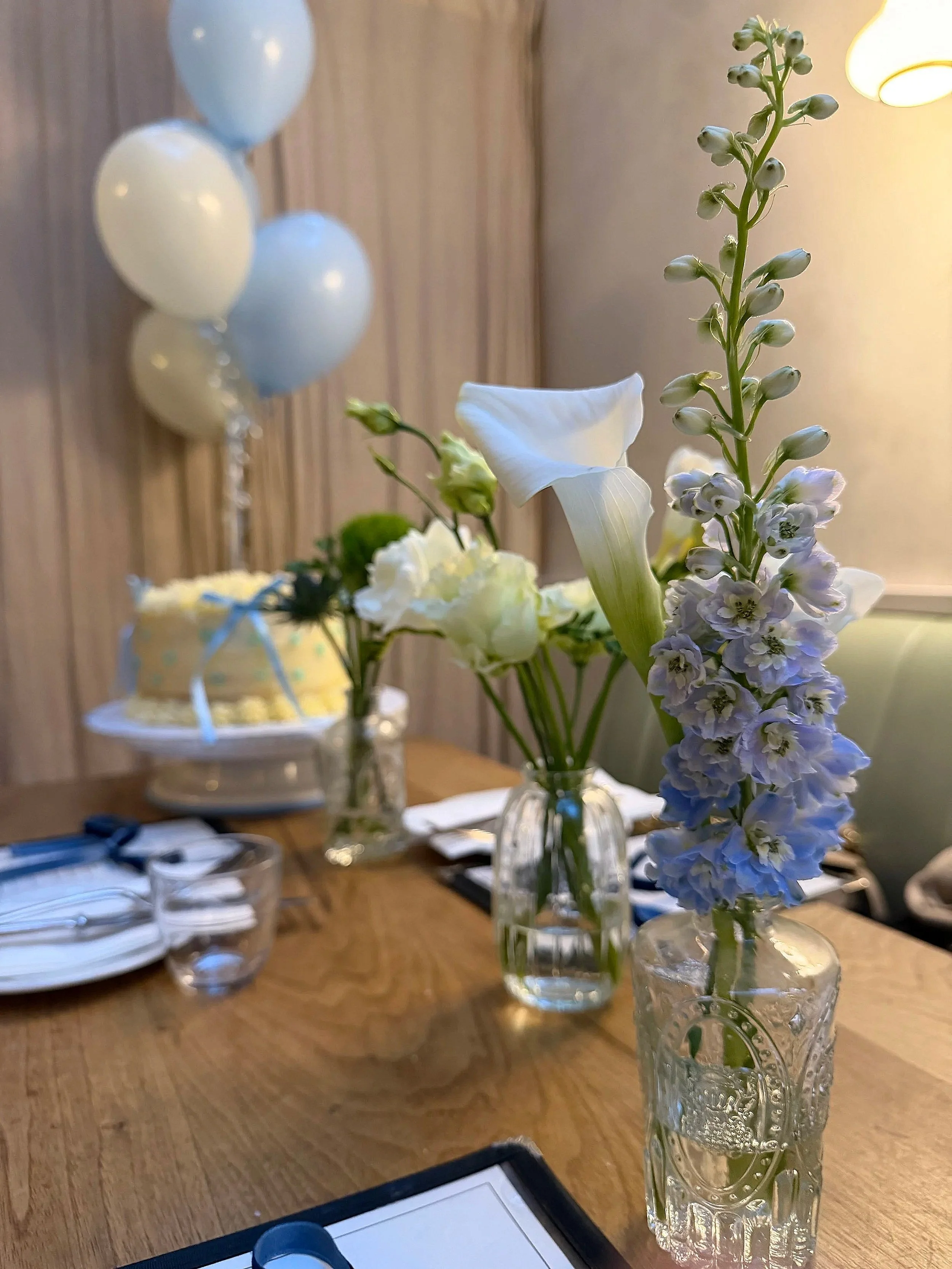 Decorative table scene with glass vases holding white and purple flowers, balloons, a cake, plates, and utensils.