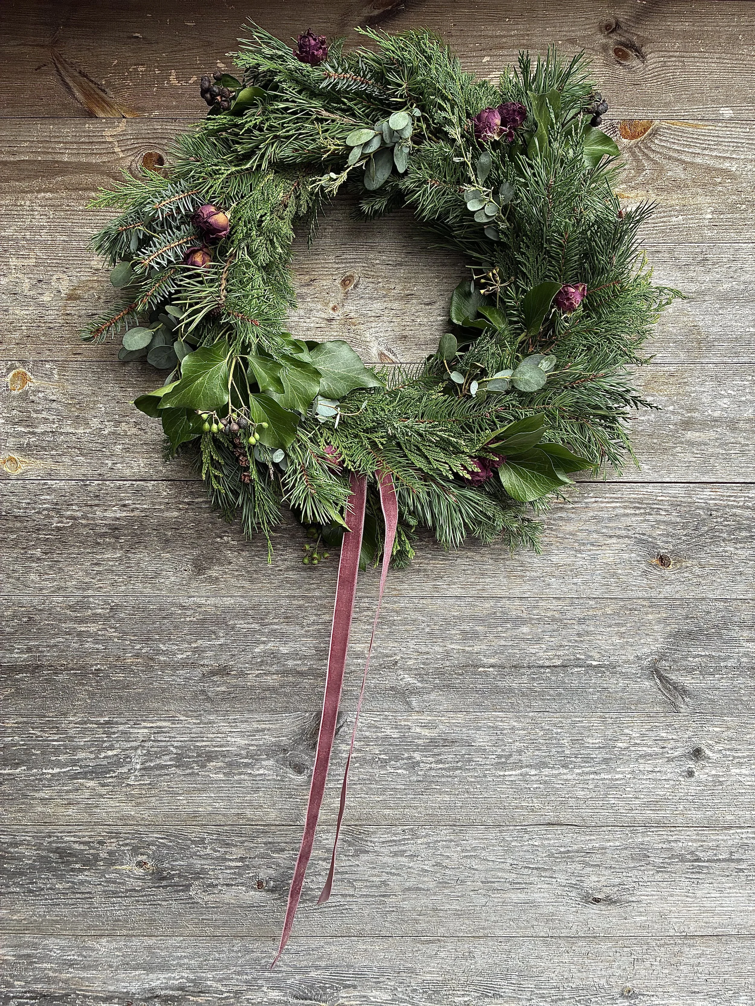A holiday wreath made of pine, holly leaves, eucalyptus, and purple flowers with a pink ribbon, hanging on a wooden surface.