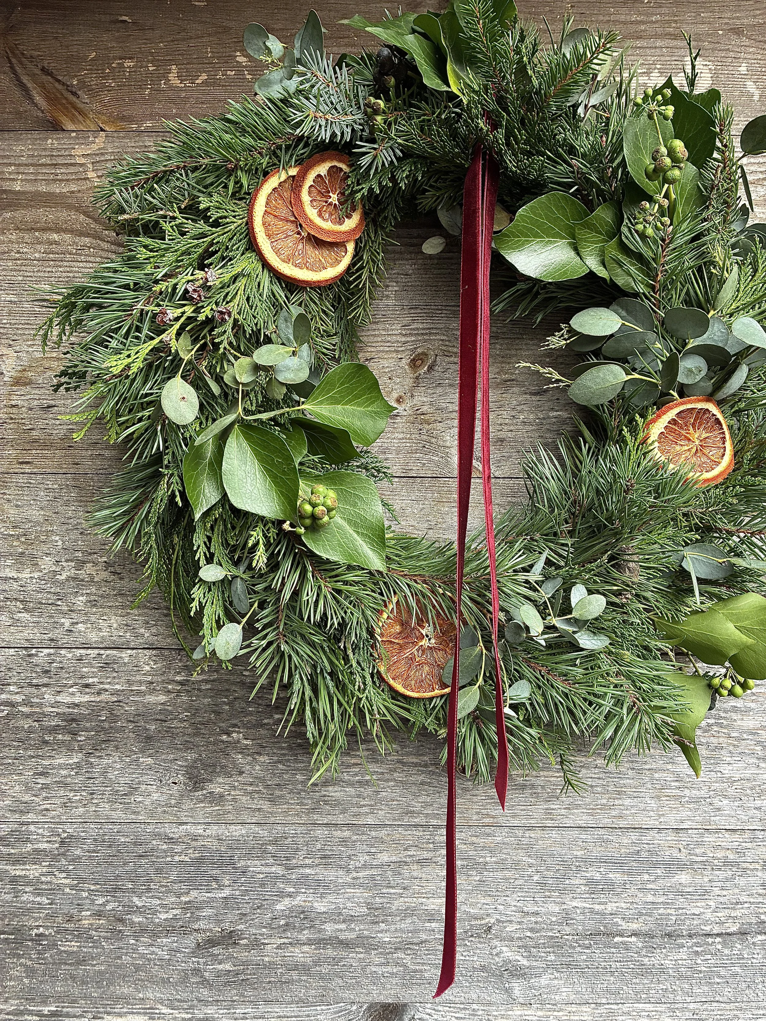 Green holiday wreath with dried orange slices and a red ribbon, hung on a wooden surface.