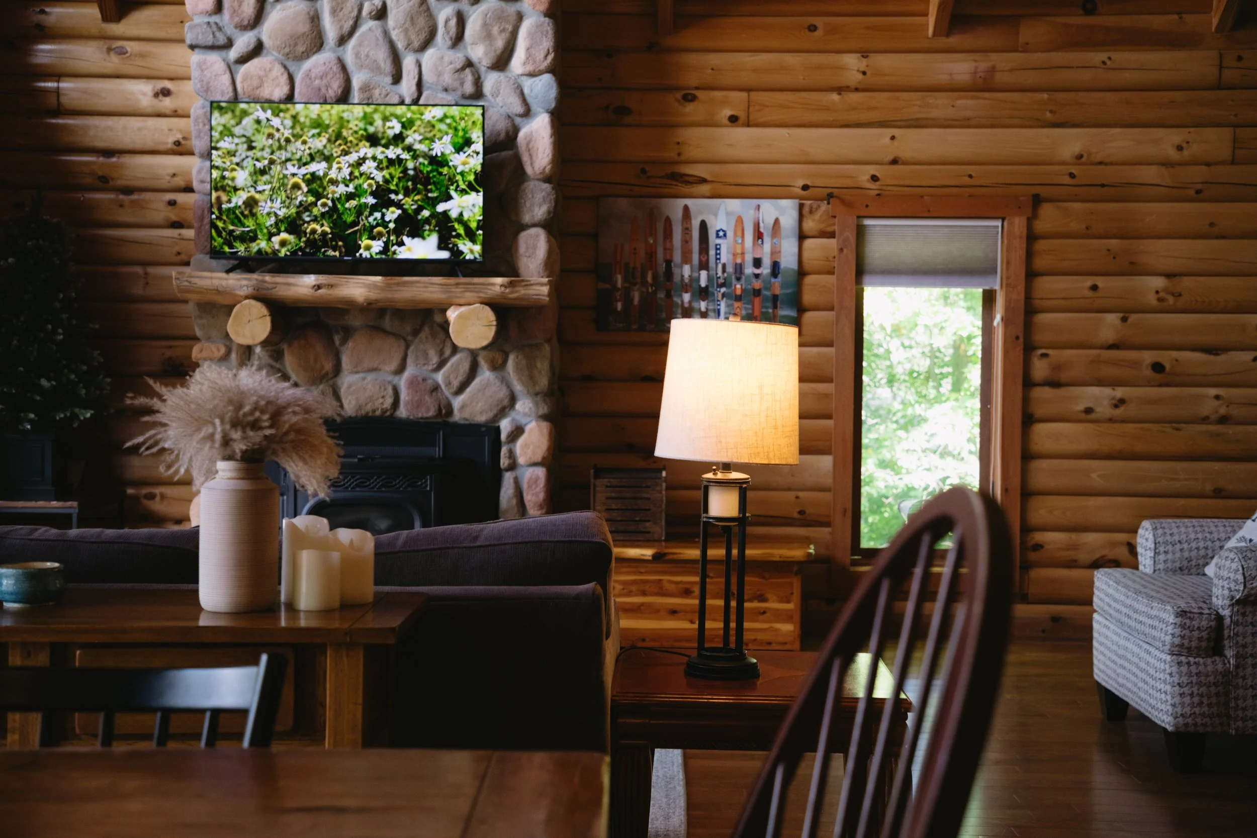 Interior of a cozy log cabin living room with wood paneled walls, a stone fireplace with a flat-screen TV above it, a wooden side table with a lamp, a potted plant, and a window showing trees outside.