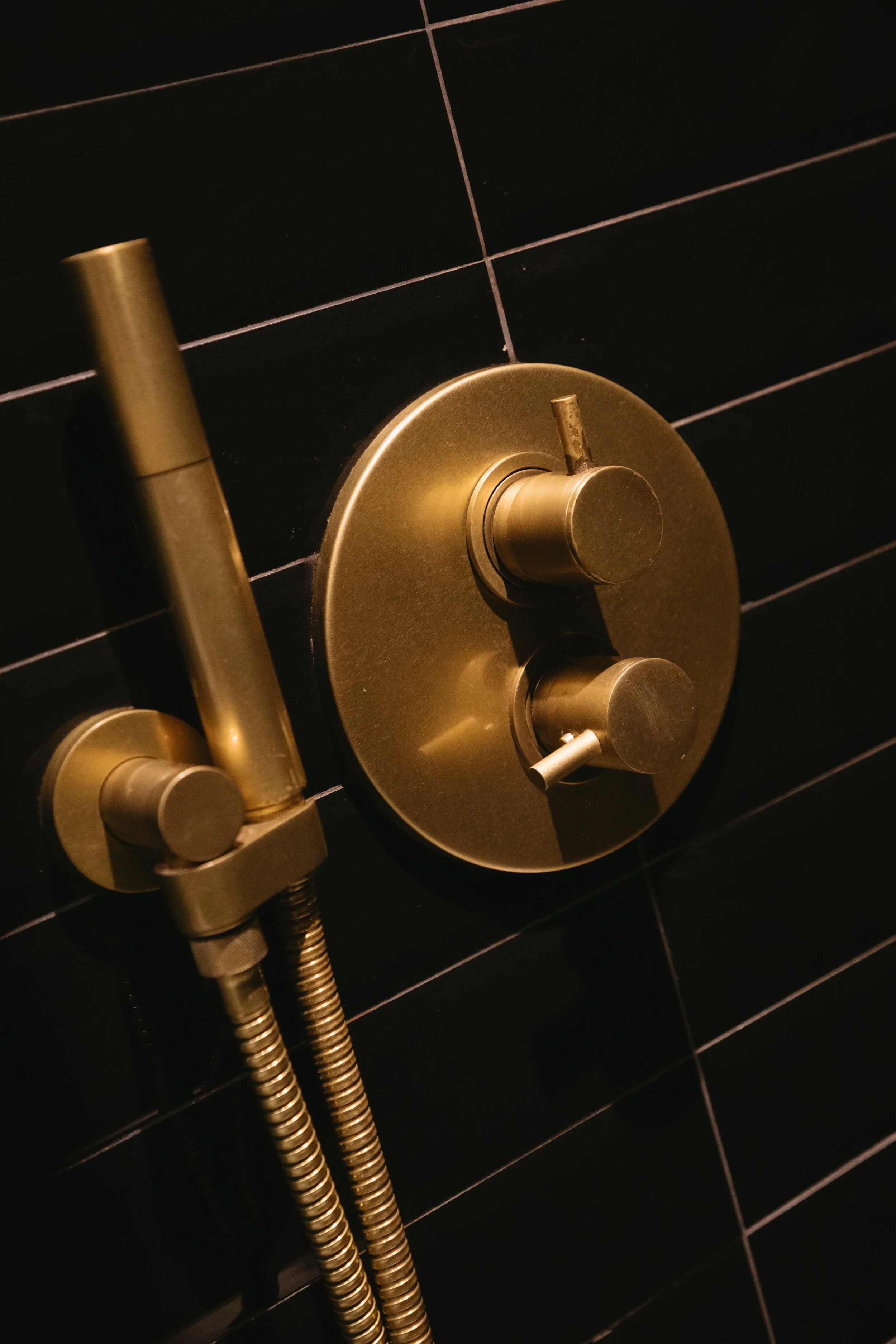 Close-up view of a modern brass shower valve and handheld shower head mounted on a black tiled bathroom wall.