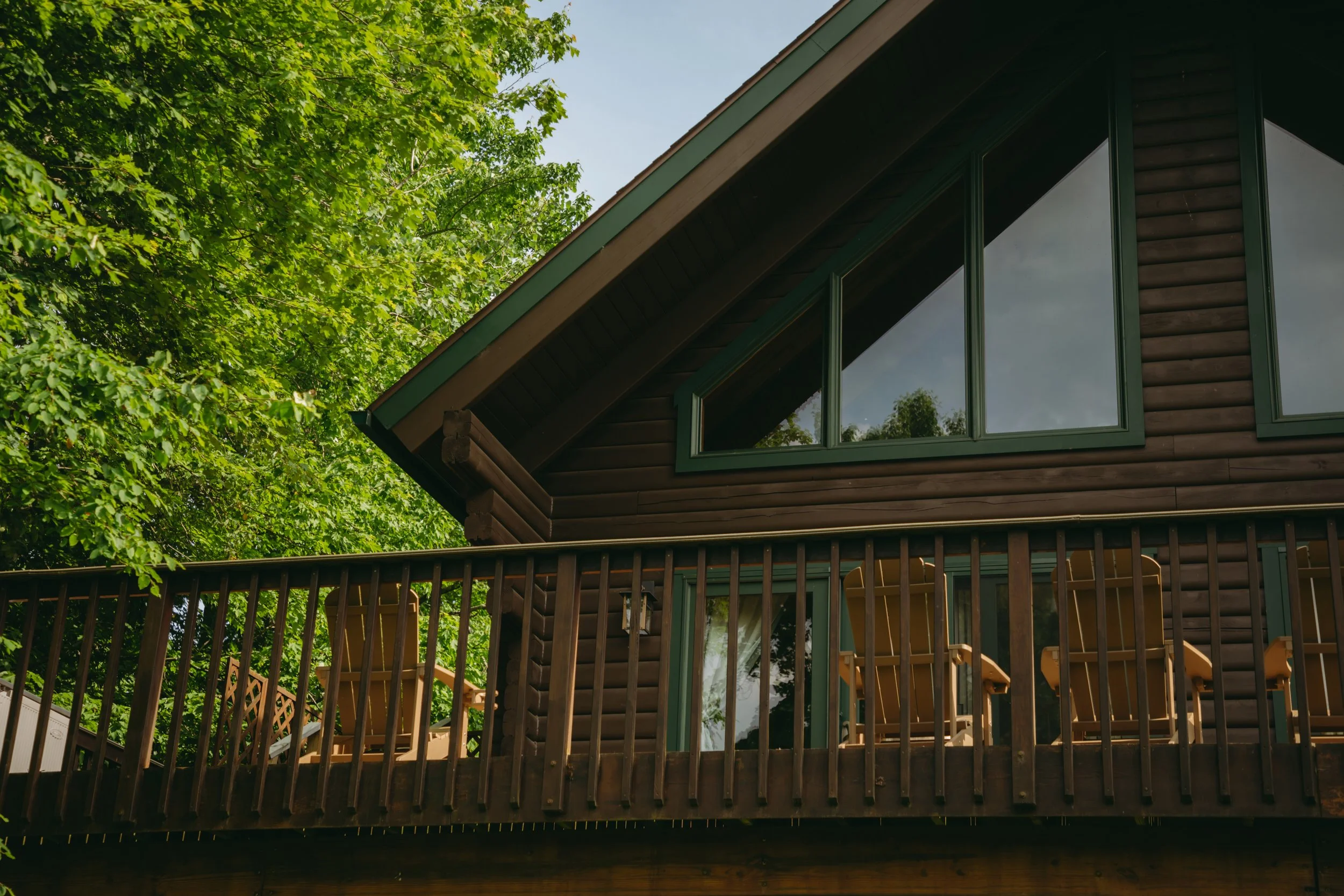 Wooden cabin with large triangular windows and a balcony with chairs, surrounded by green trees.