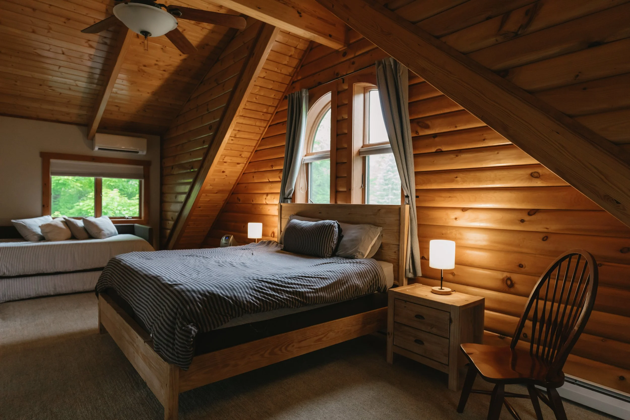 A cozy bedroom with wood-paneled walls and sloped ceiling, featuring a wooden bed with striped bedding, a nightstand with a lamp, a large window with curtains, a seating area by the window, and a wooden chair.