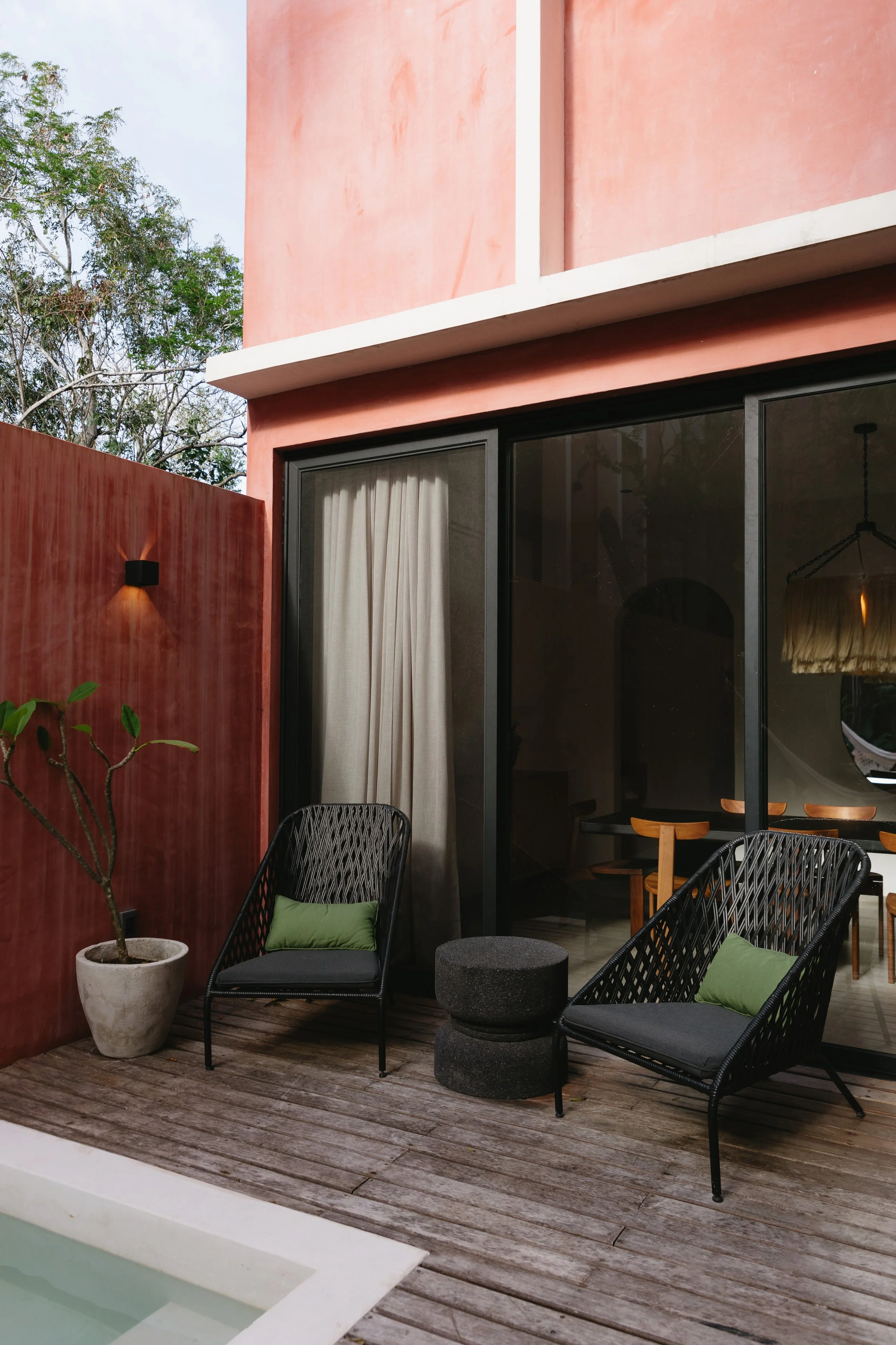 Outdoor patio with two black woven chairs with green pillows, a small black side table, potted plant, and a pool, adjacent to a pink house with large sliding glass doors.