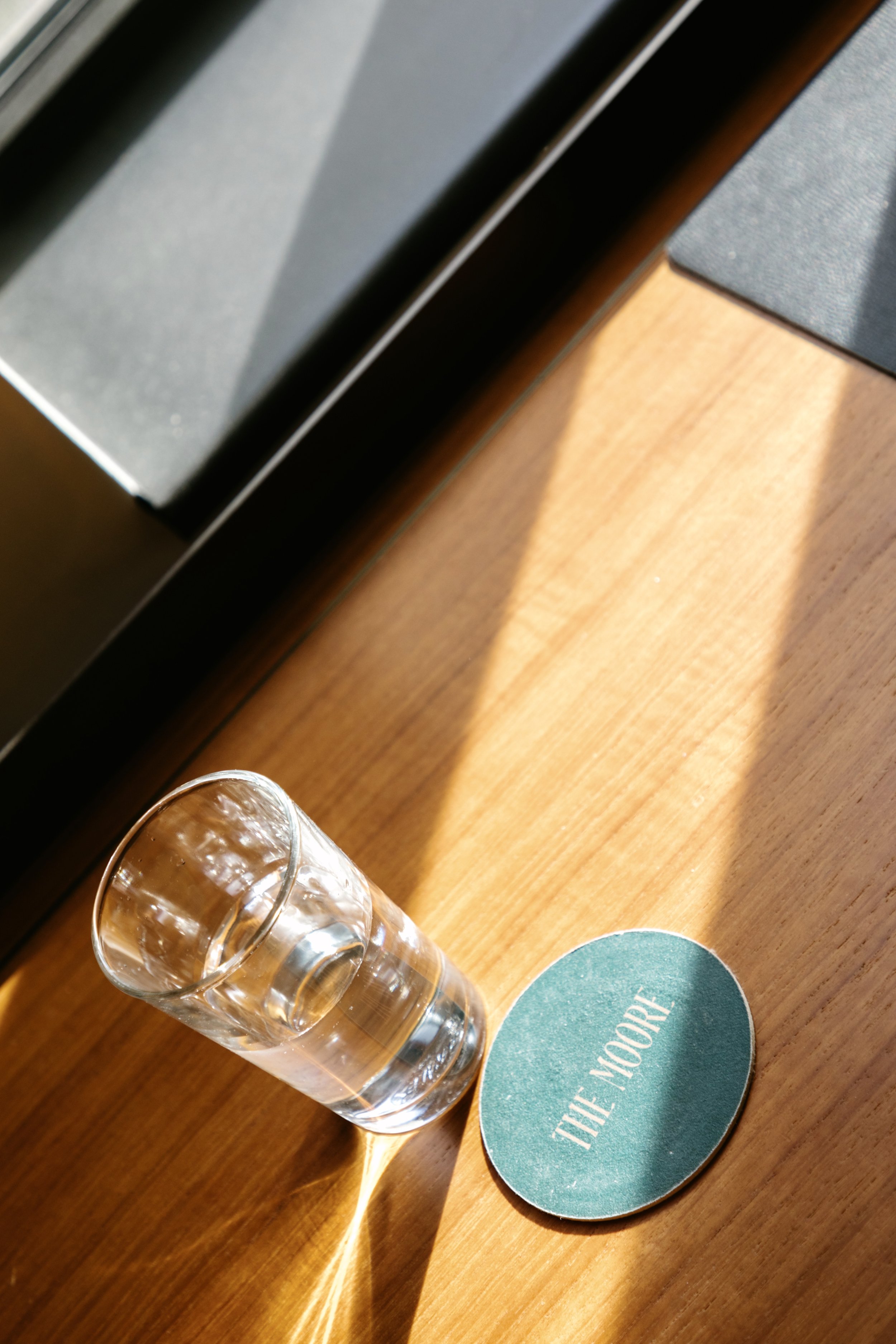 A glass of water on a wooden surface next to a teal paper coaster with the words 'THE MOORE' on it, with sunlight casting shadows.