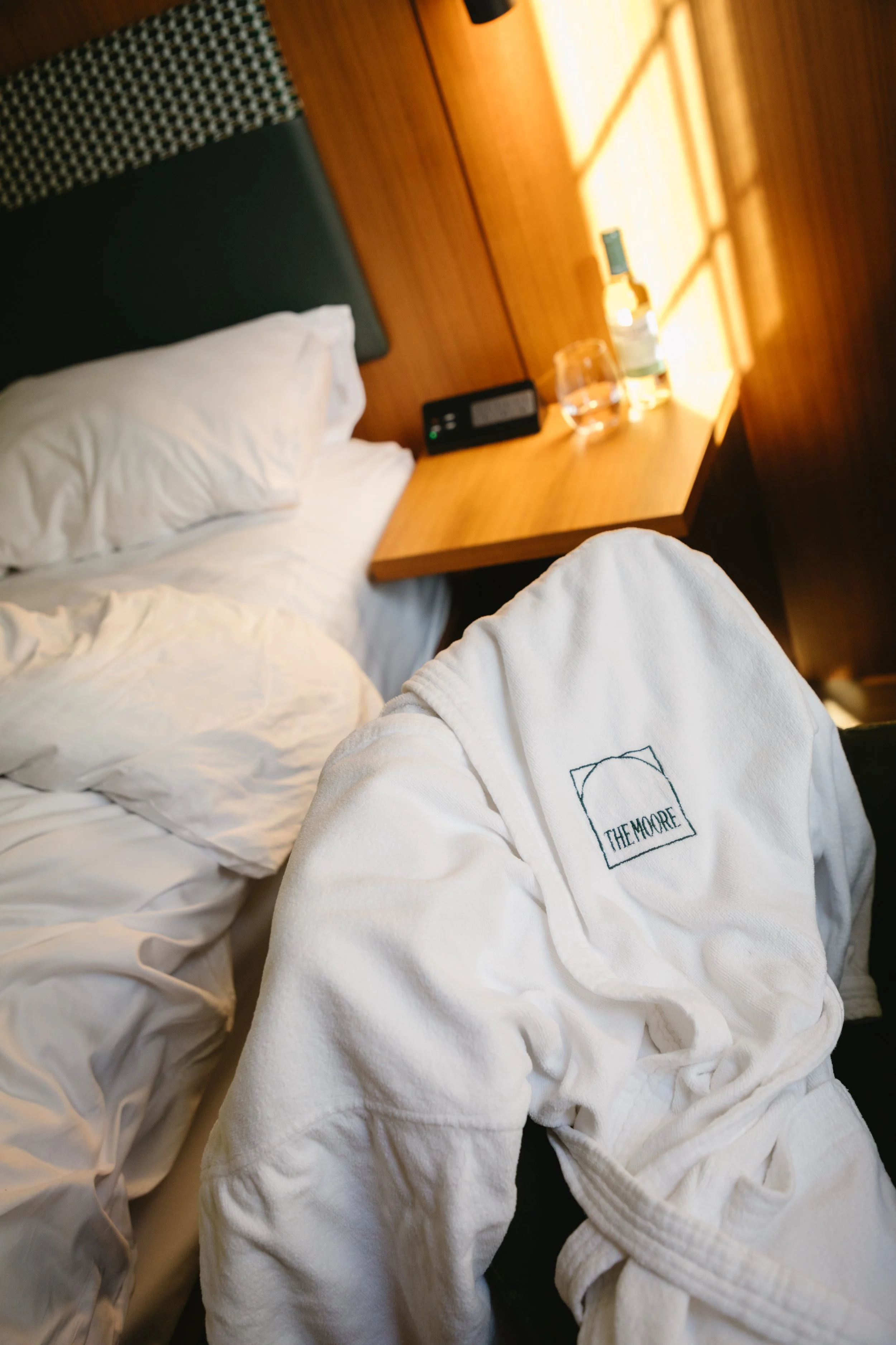 Hotel room with a bed, nightstand, and a person in a white bathrobe with a logo that reads 'THE MOORE'.