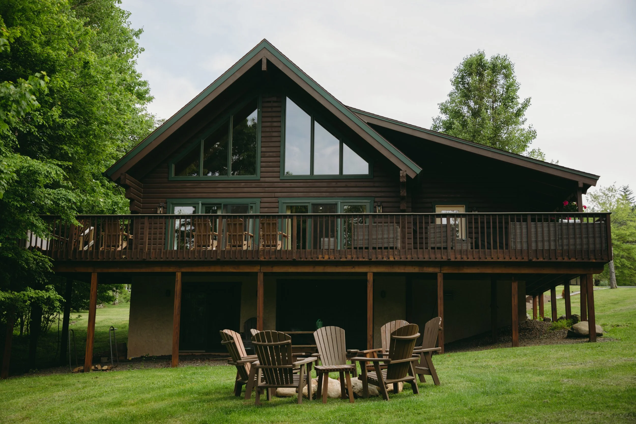 A multi-story wooden house with large triangular windows and a spacious balcony, surrounded by green trees and a well-kept lawn with outdoor chairs and a small table.