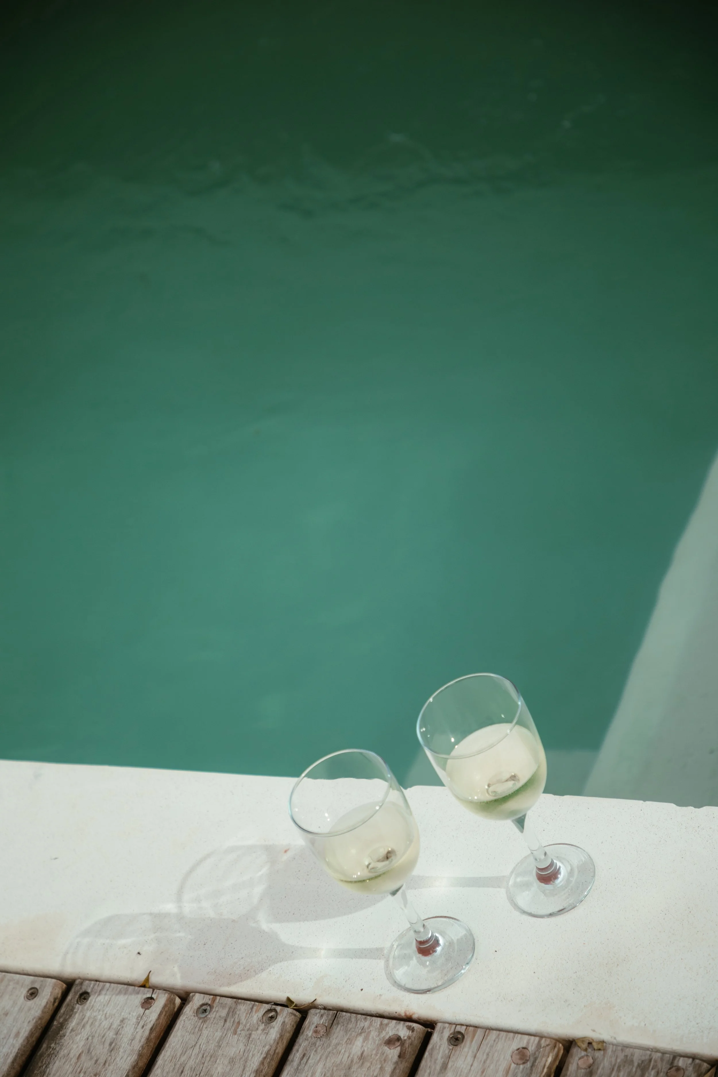 Two glasses of white wine placed on a white poolside ledge, with a swimming pool in the background.