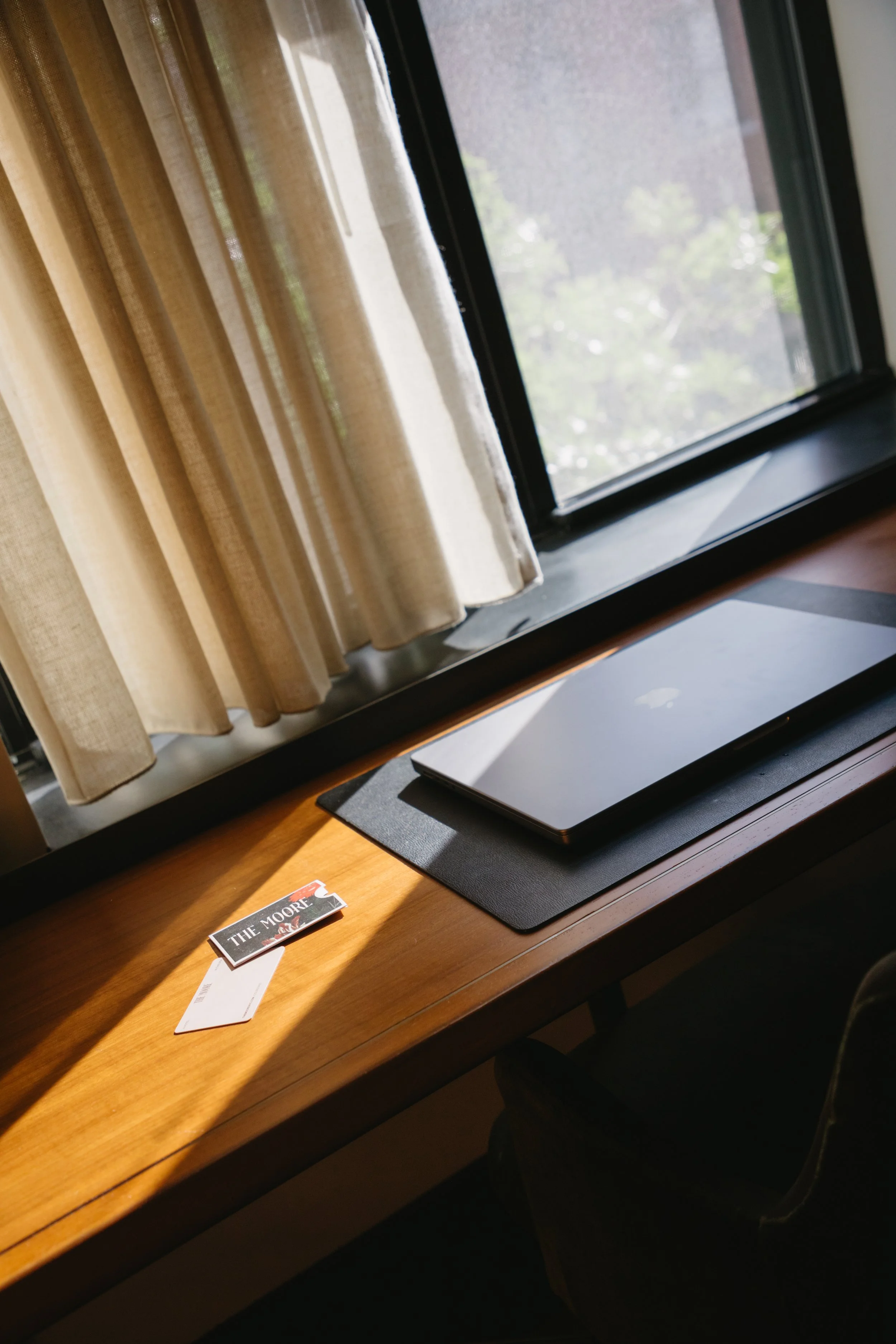 Wooden desk near a window with a laptop on a black mousepad, curtains, and a couple of cards.