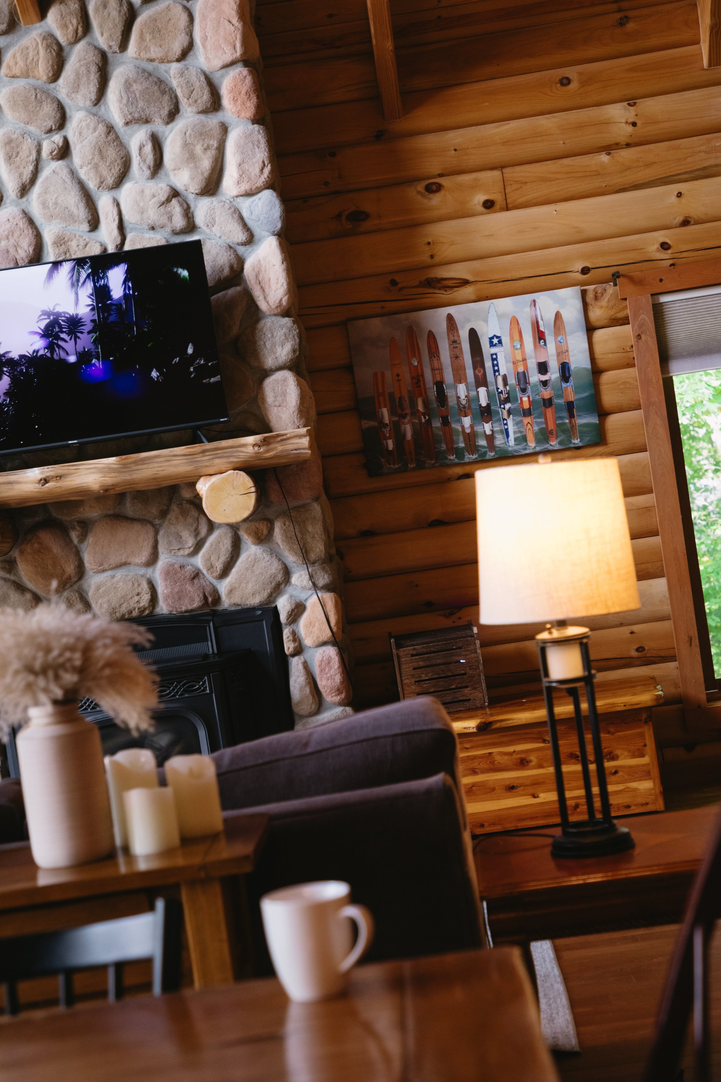 Living room with wooden walls, stone fireplace, wall-mounted TV, lit table lamp, and wall art of vintage skis.