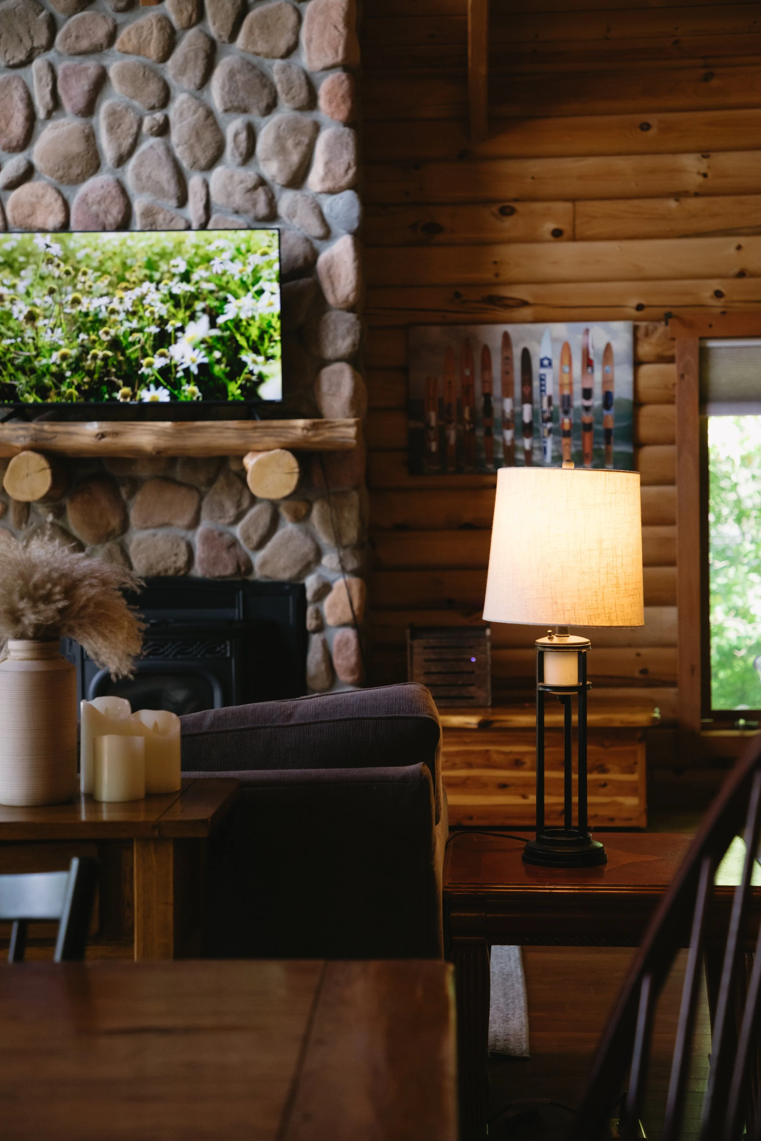 Cozy living room with stone and wood walls, a television showing flowers, a table lamp, a painting with sticks, and various decorations, illuminated by natural light.