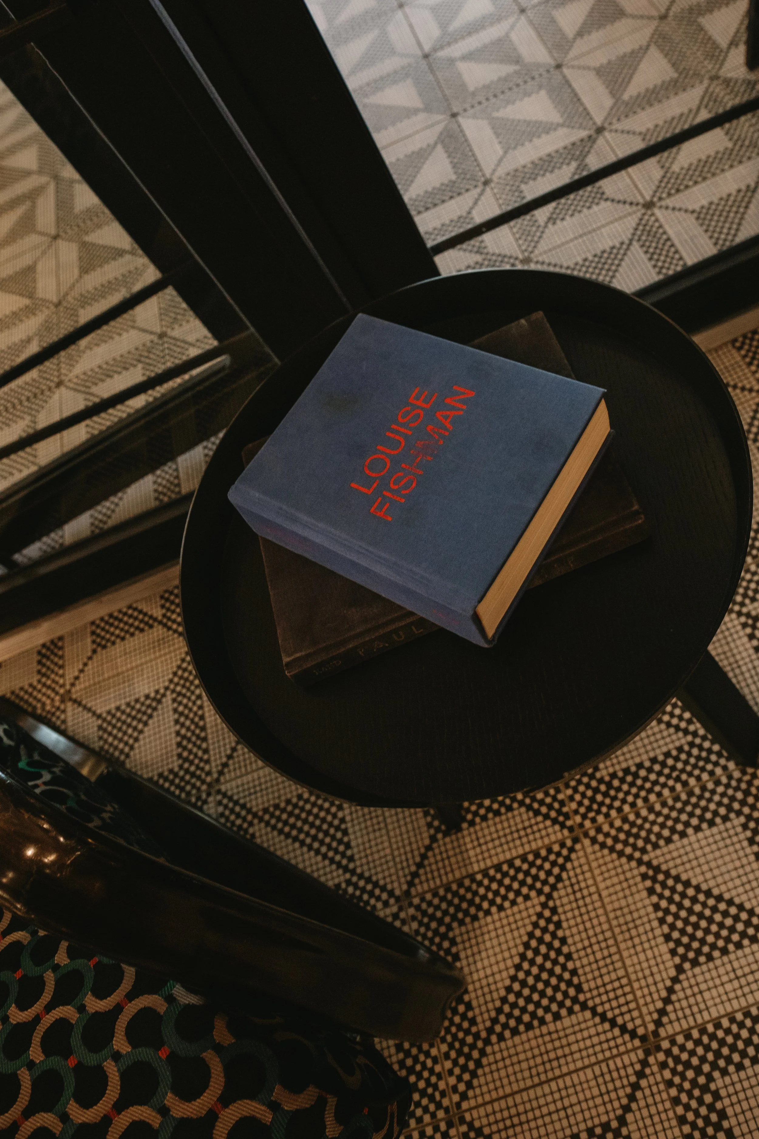 A round black table holding three books, with the top one titled 'Louis-Frederic Felschmann', placed next to a chair and a floor with black and white geometric patterned tiles.