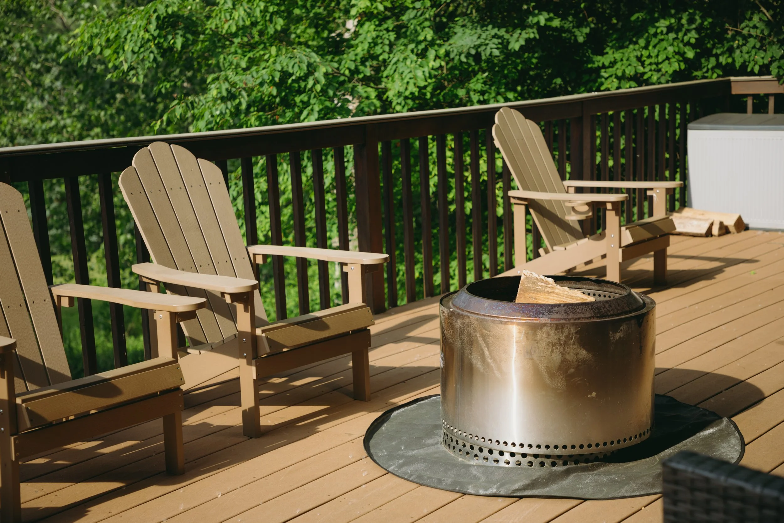 A wooden deck with Adirondack chairs, a fire pit with wood inside, and a white storage box, with green trees in the background.