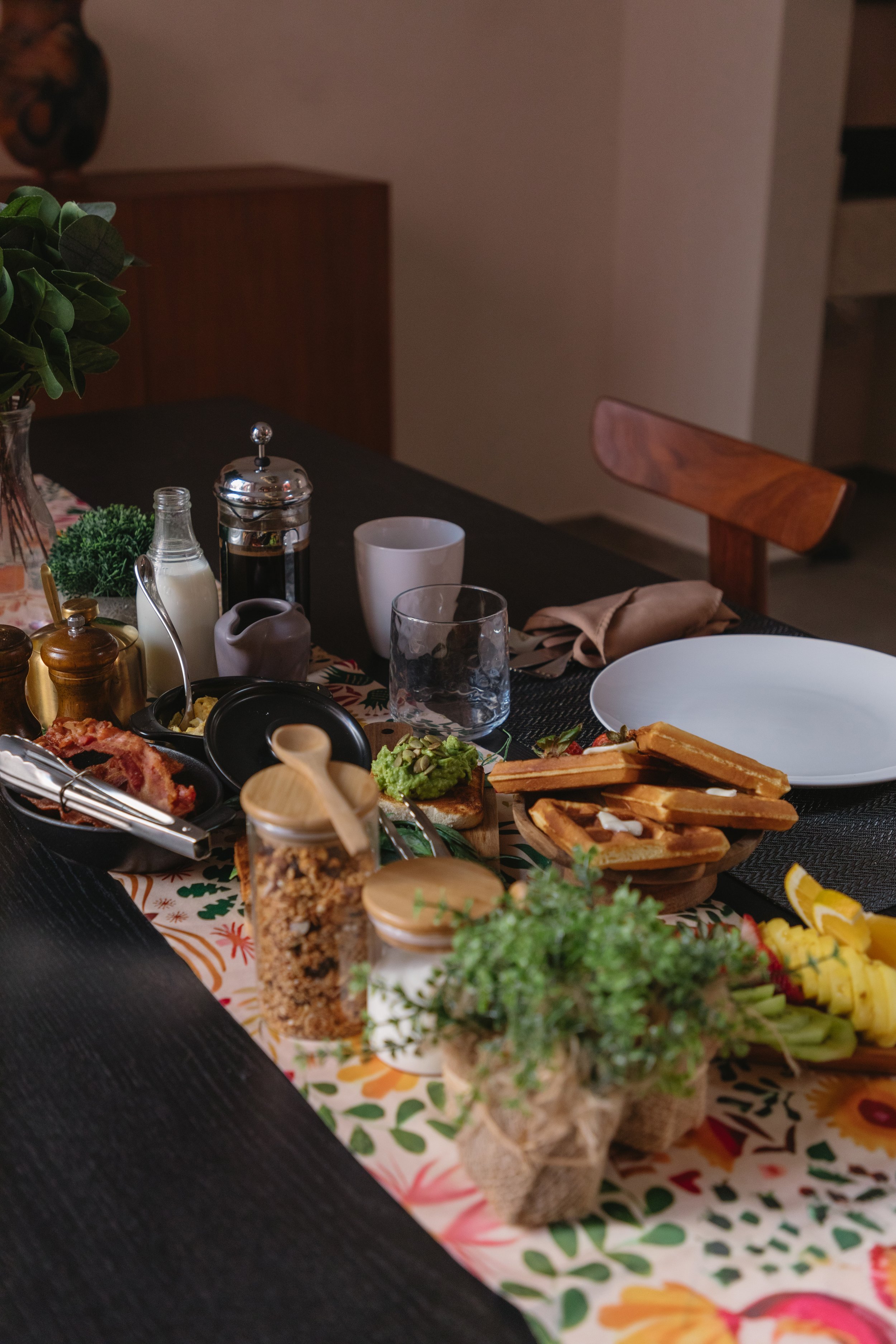 A table set with various breakfast foods including toast with spread, pineapple slices, a bowl of guacamole with vegetables, jars of granola and honey, a water glass, mugs, and condiments, all on a colorful table runner.