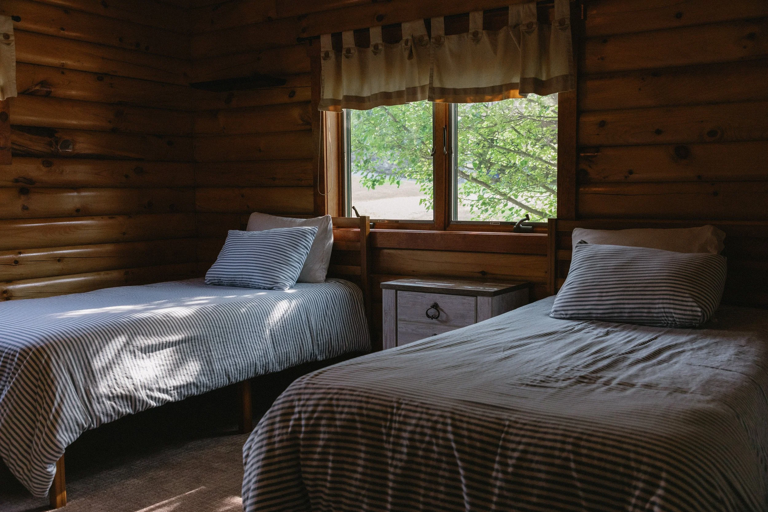 Two twin beds with striped bedding and pillows in a rustic log cabin bedroom, with a window showing green tree branches outside.