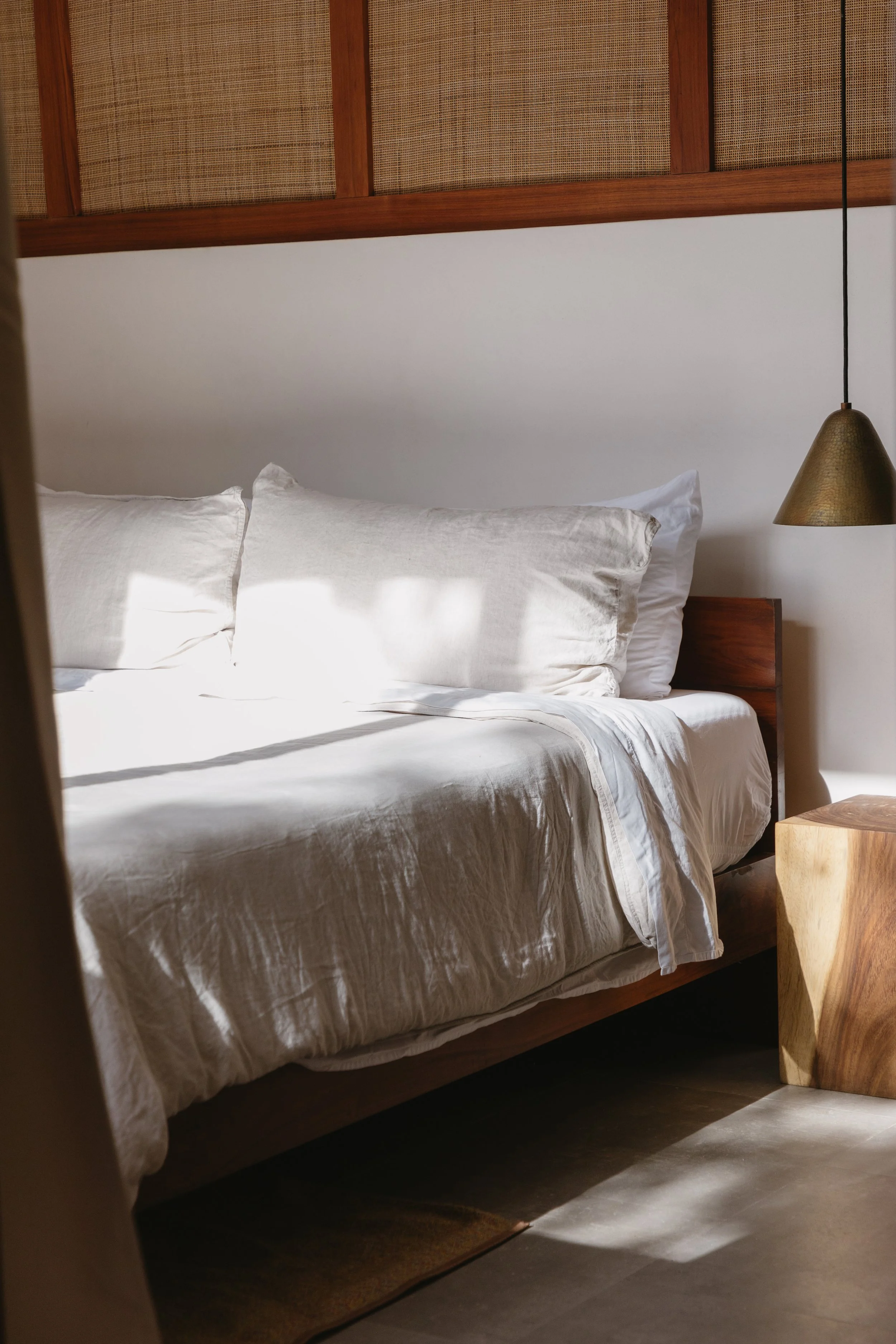 A neatly made bed with white linens and pillows, a wooden headboard, and a modern wooden bedside table with a hanging gold-colored lamp.