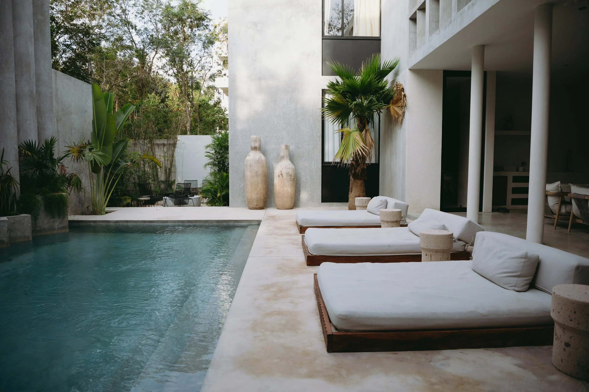 Modern outdoor pool area with white lounge chairs and large potted plants, adjacent to a contemporary building with white walls and large vases.