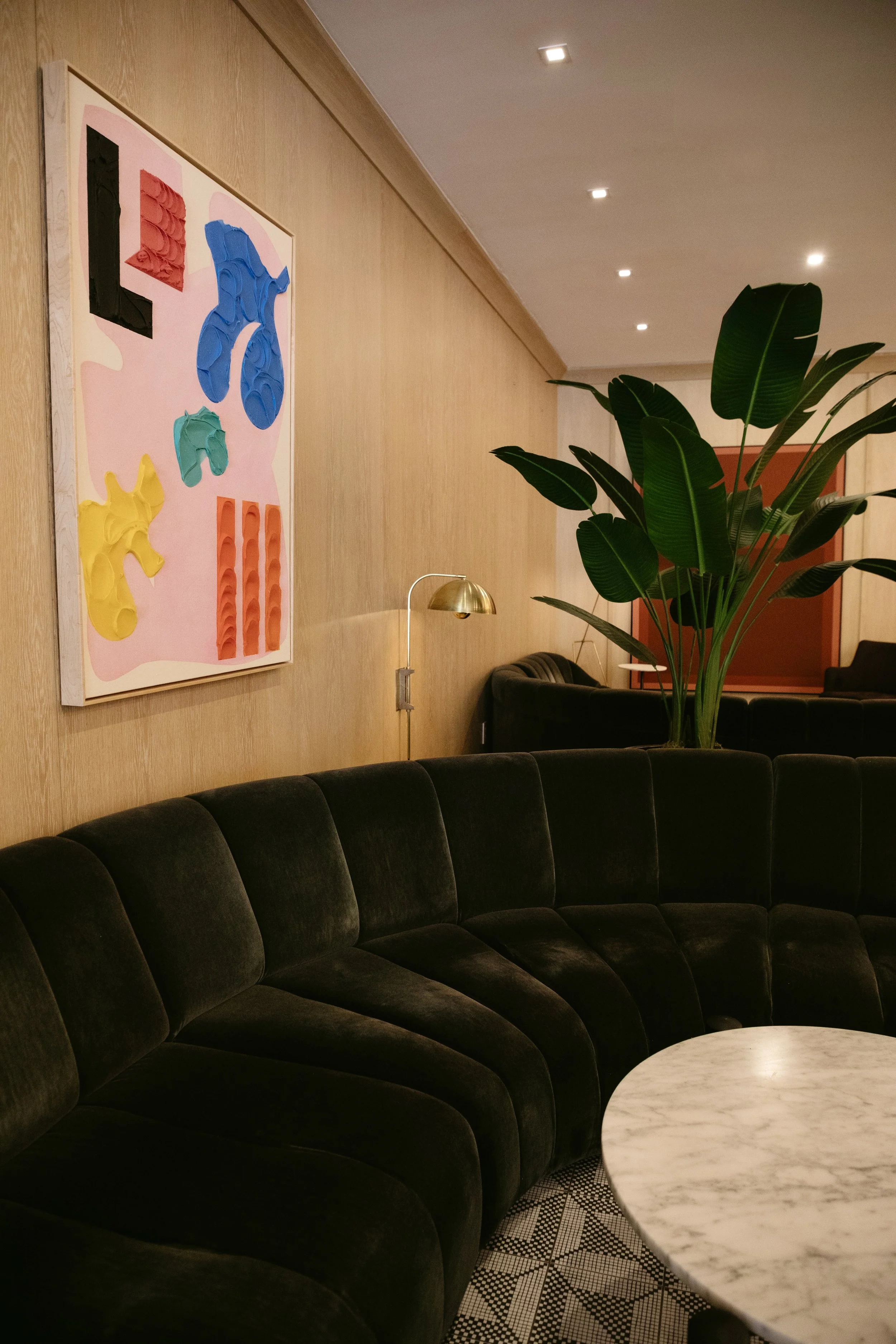 A modern hotel lobby with a curved black velvet sofa, a white marble round table, a large green plant, wooden wall paneling, a colorful abstract artwork, a brass floor lamp, and recessed ceiling lights.