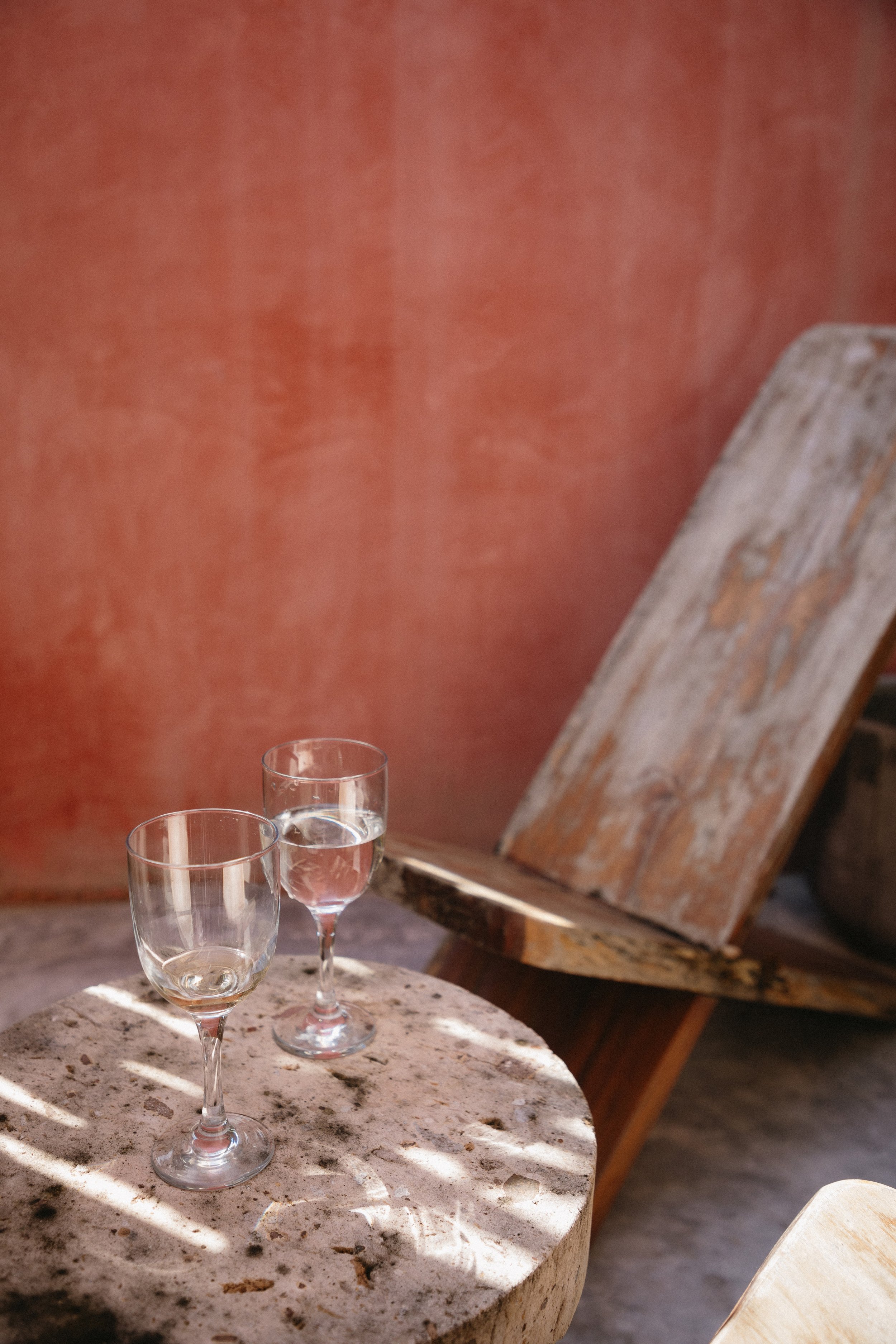 Two glasses of water on a stone table with a rustic wooden chair and a textured warm-colored wall in the background.