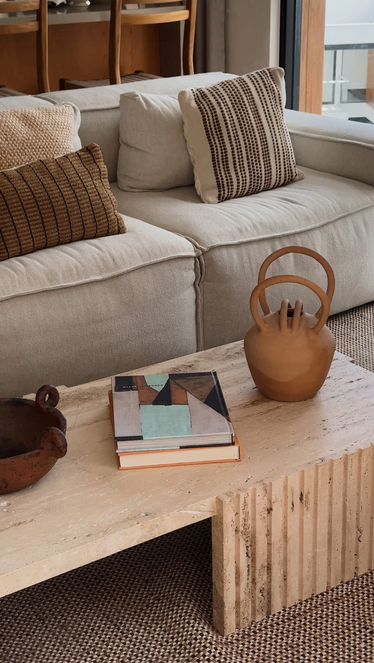 A beige sofa with three patterned throw pillows, a wooden coffee table with decorative items and books, and a window in the background.