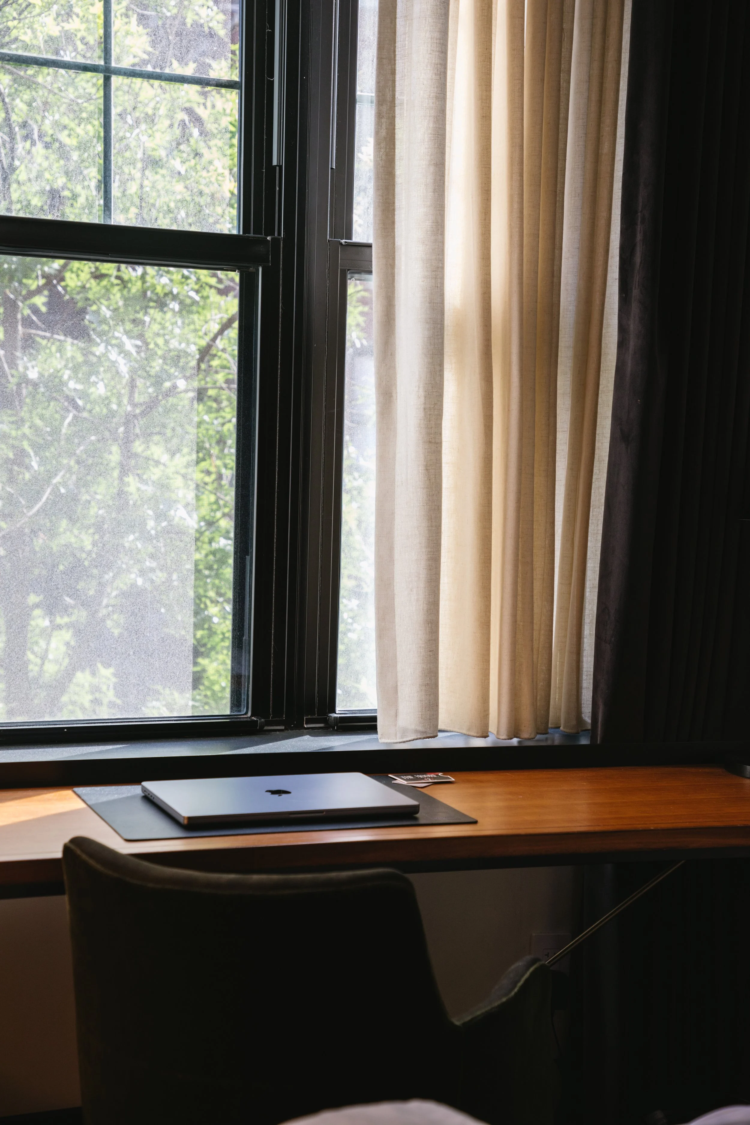 A wooden desk with a closed silver laptop placed on a mat, positioned in front of a large window with black frames. The window has partially open cream-colored curtains, revealing green trees outside.