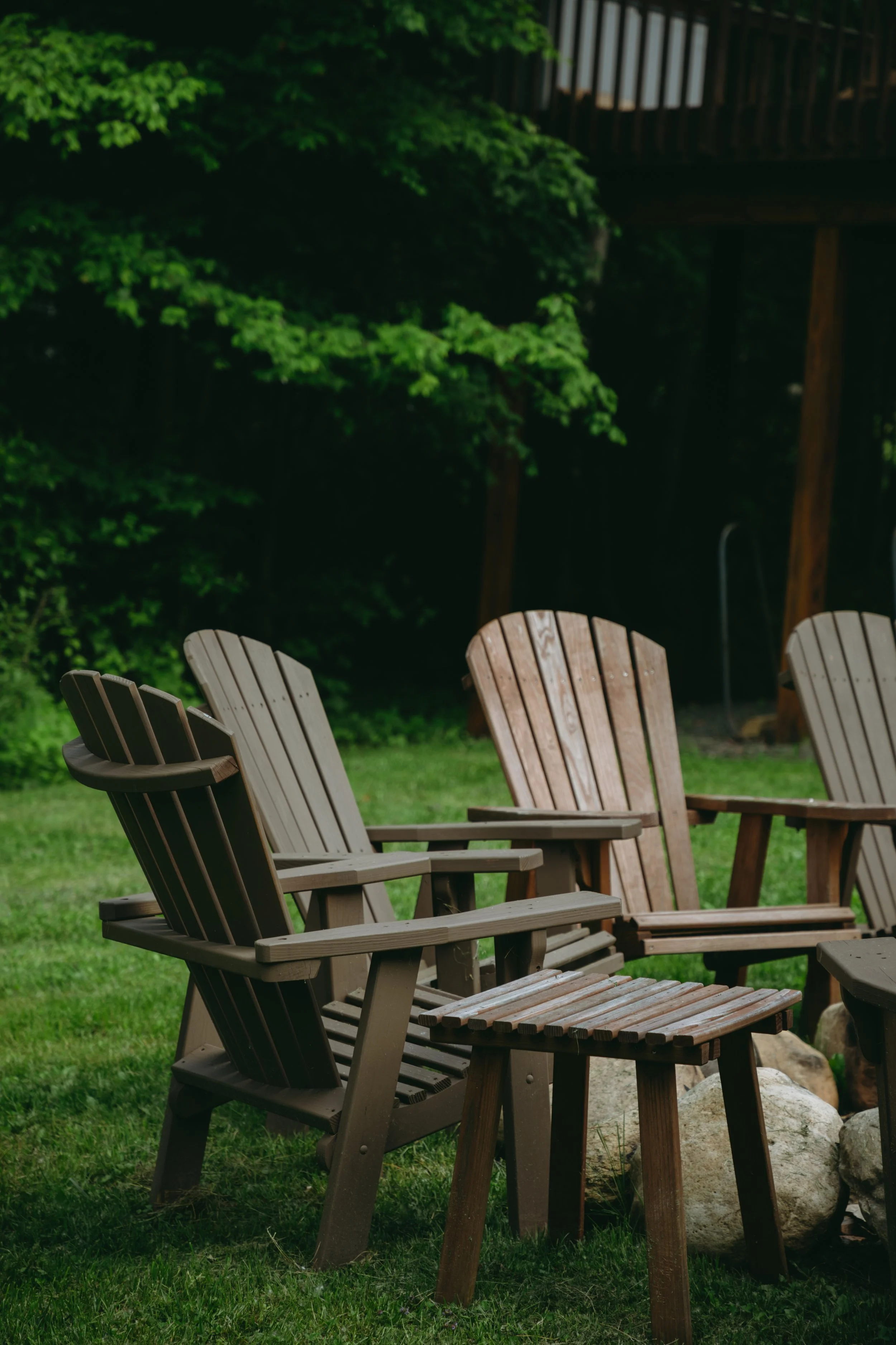 Several Adirondack chairs and small table outside on grass, with trees and a wooden railing in the background.